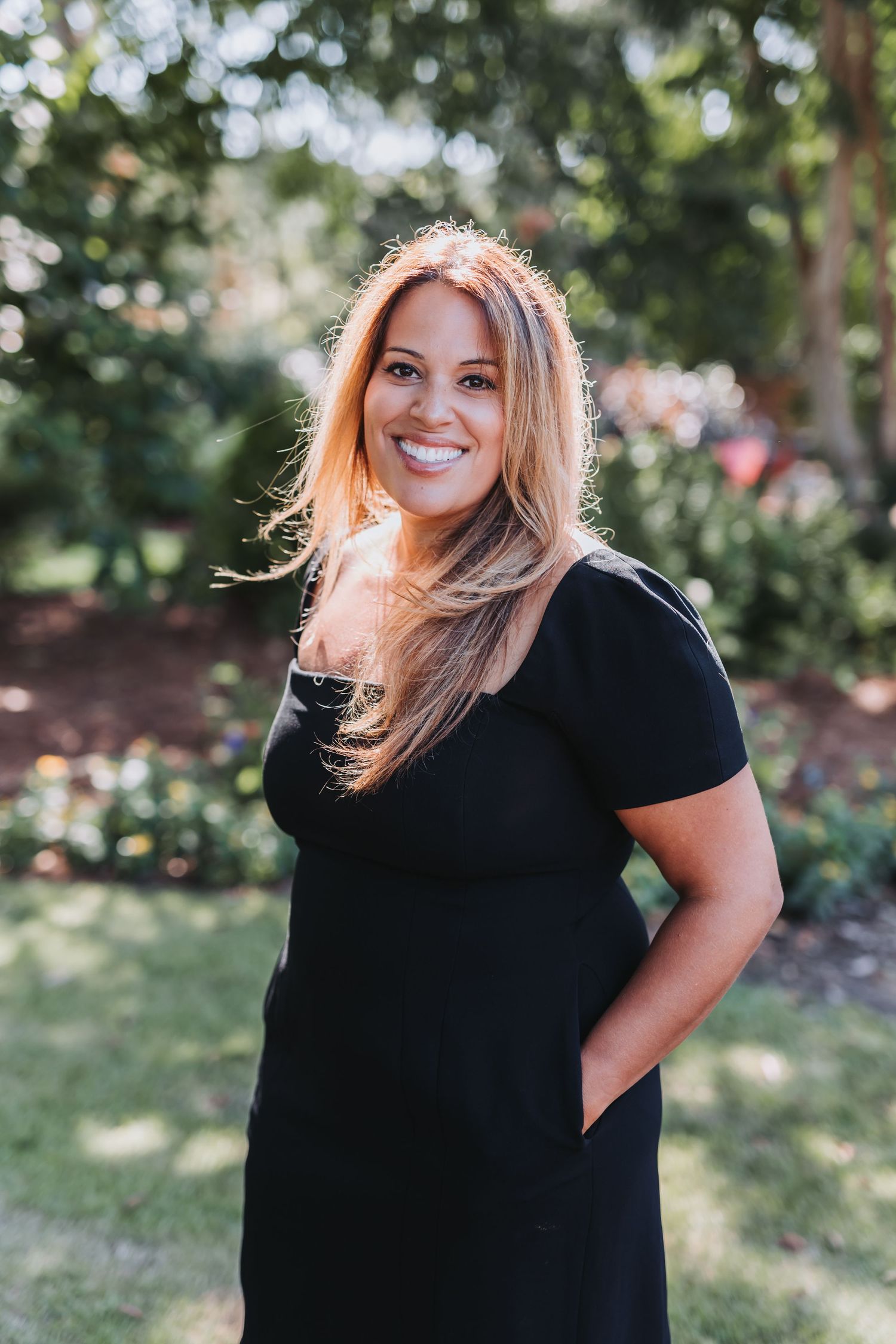Smiling woman with long blonde hair wearing a black dress poses in a lush green garden setting.