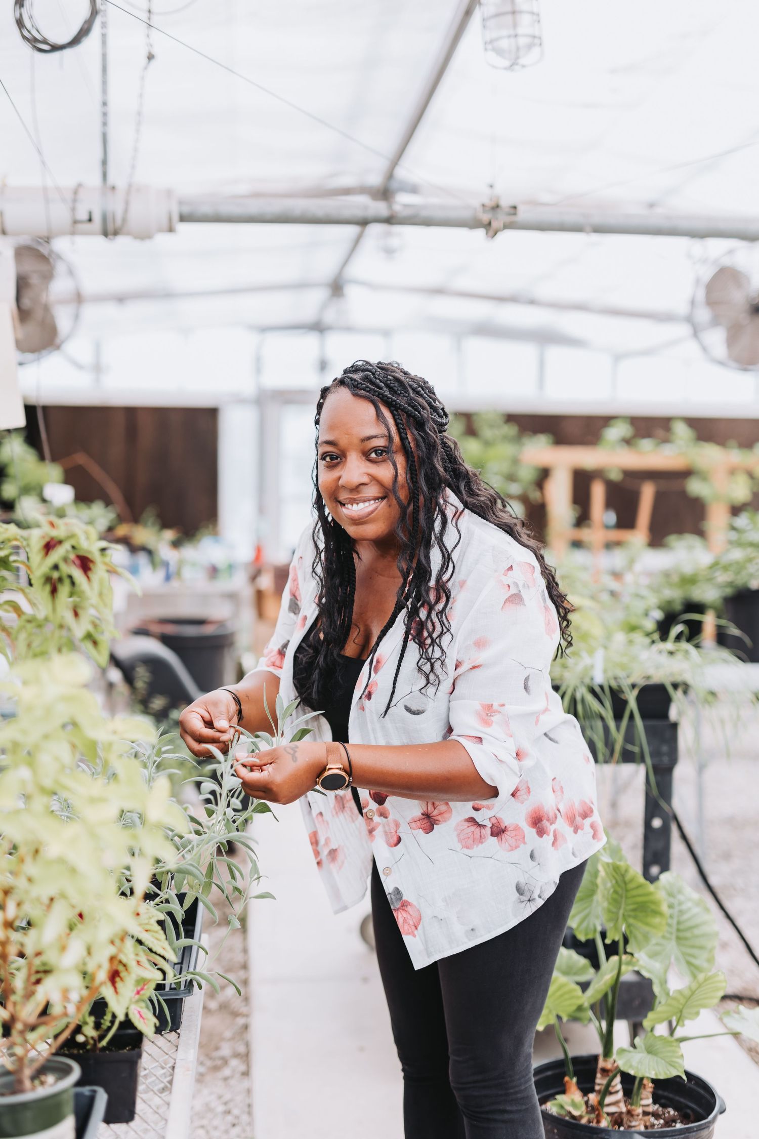 Smiling woman in floral shirt tending to plants in a greenhouse nursery.