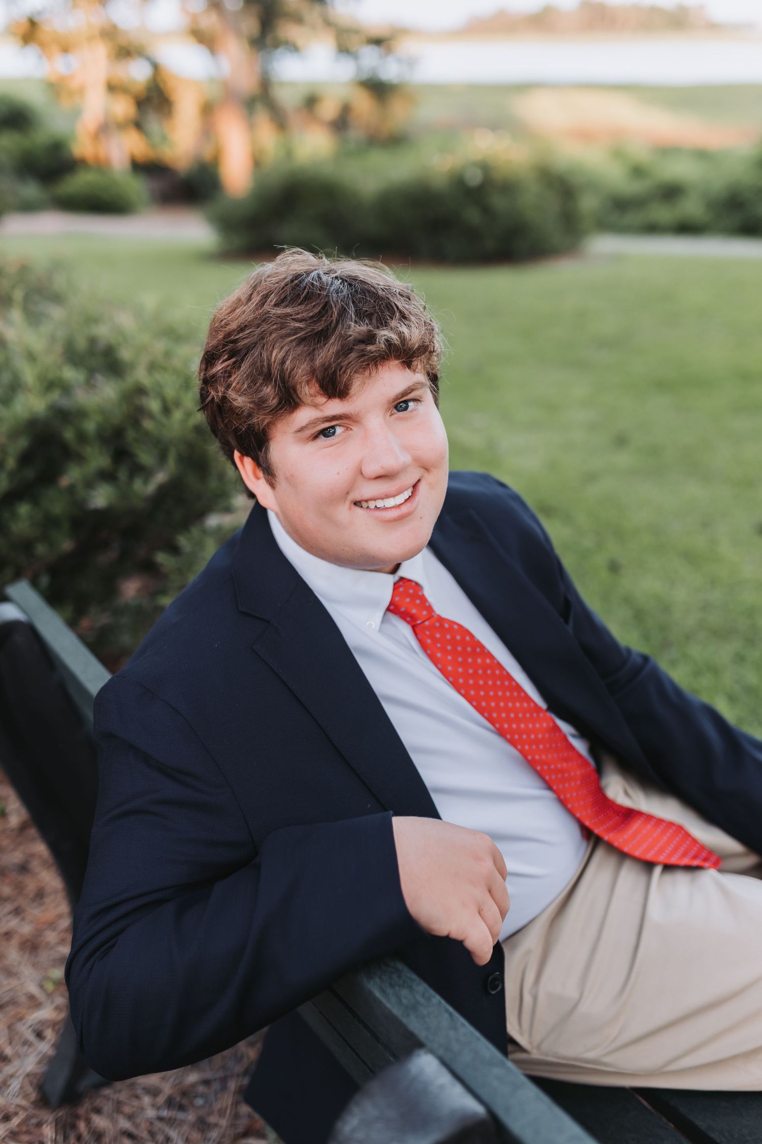 Young man in navy blazer and red tie smiling on a bench in a lush green park setting.