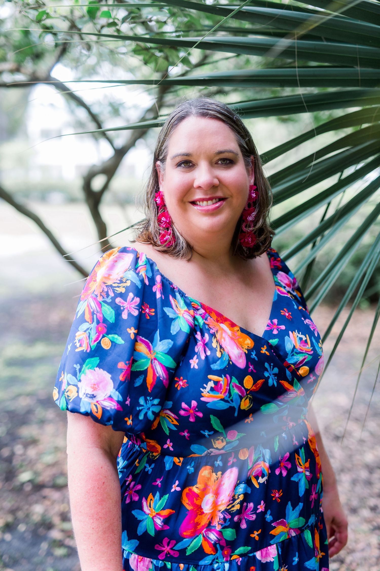 Woman smiling in colorful floral blue dress with pink earrings, standing outdoors near tropical palm leaves.