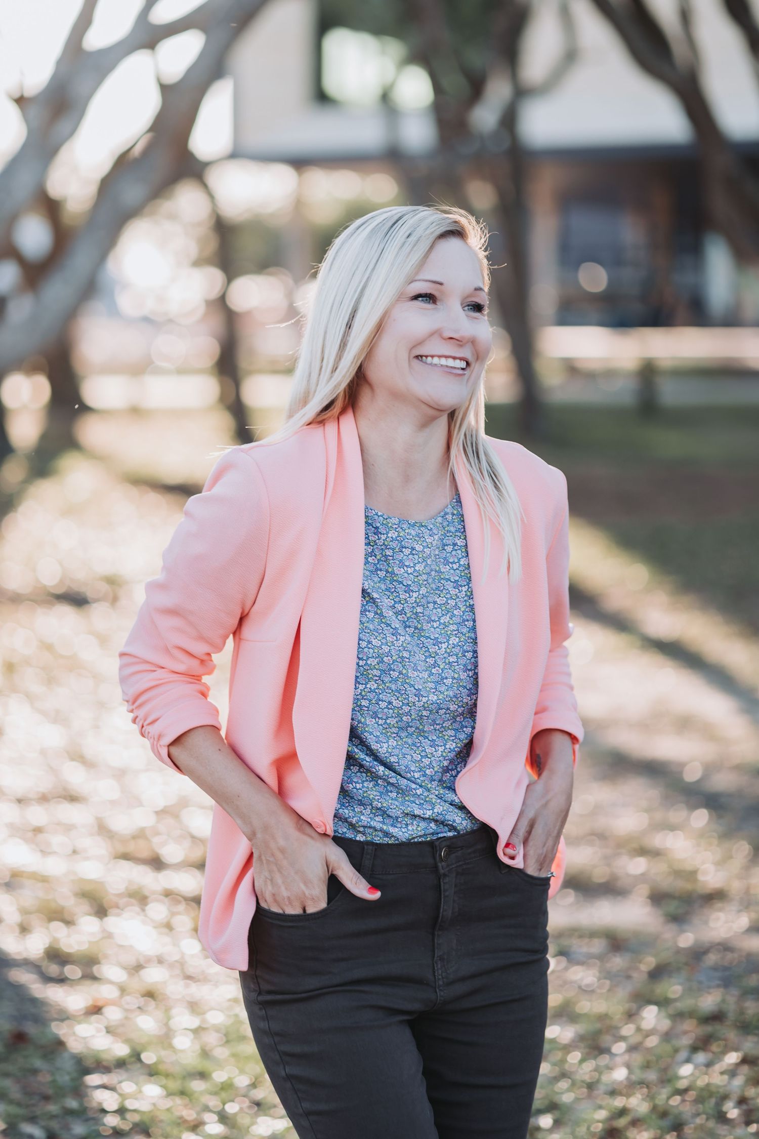 Smiling blonde woman in pink blazer and gray floral top with hands in pockets outdoors.