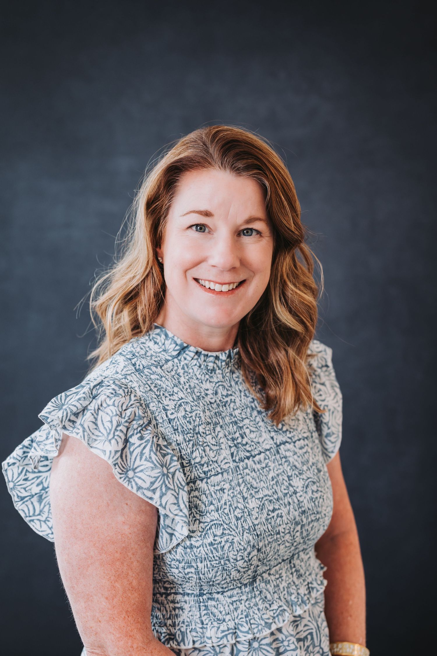 Smiling woman with auburn hair wearing a floral ruffle-sleeve blouse poses against a dark gray background.