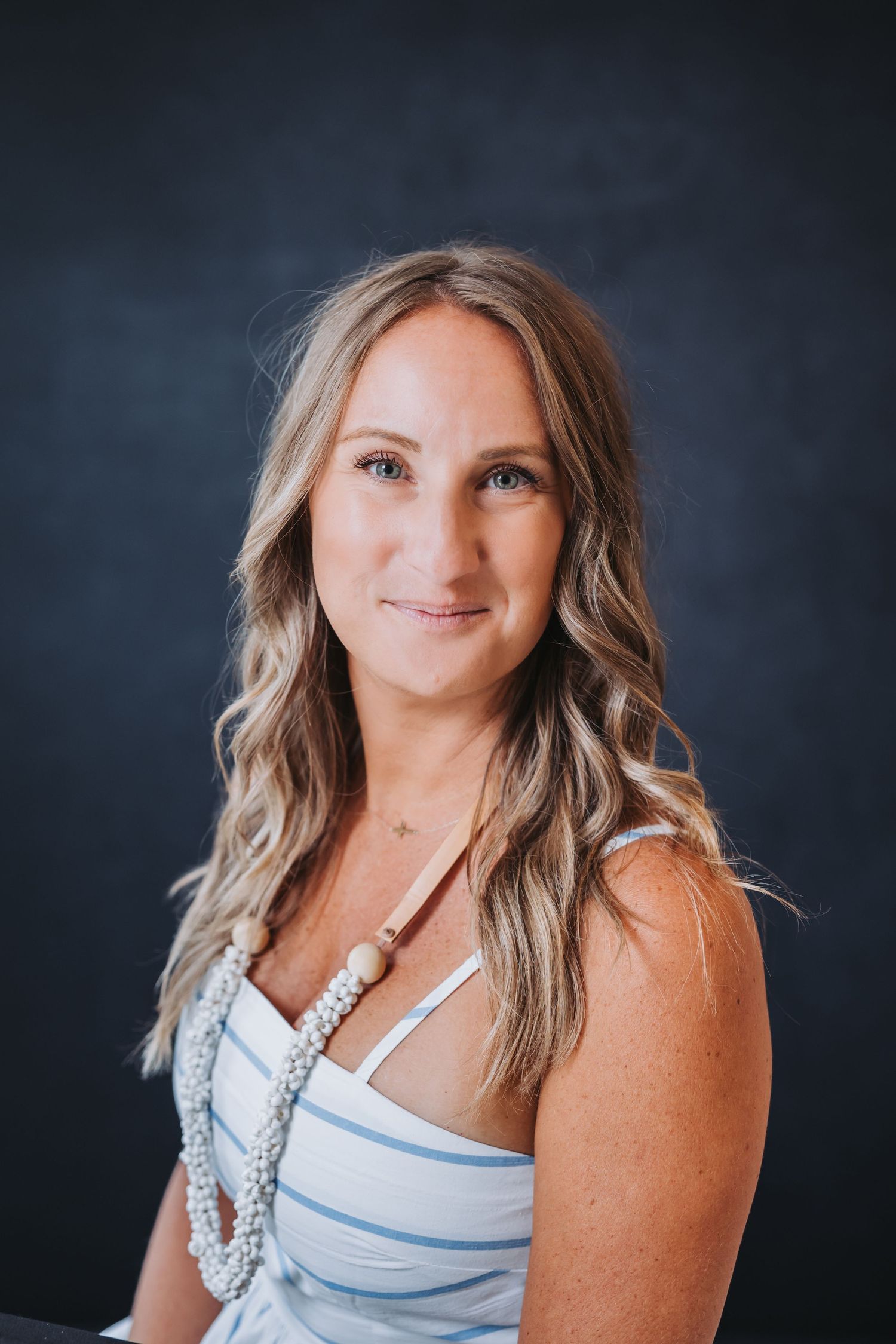 Smiling woman with blonde wavy hair wearing a striped dress and beaded necklace against a dark background.