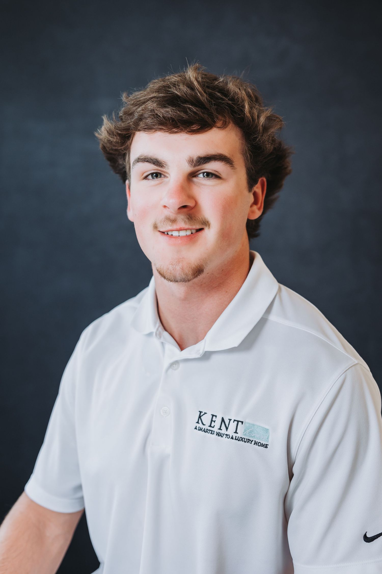Young man smiling in white Kent polo shirt against dark background, professional headshot photo.