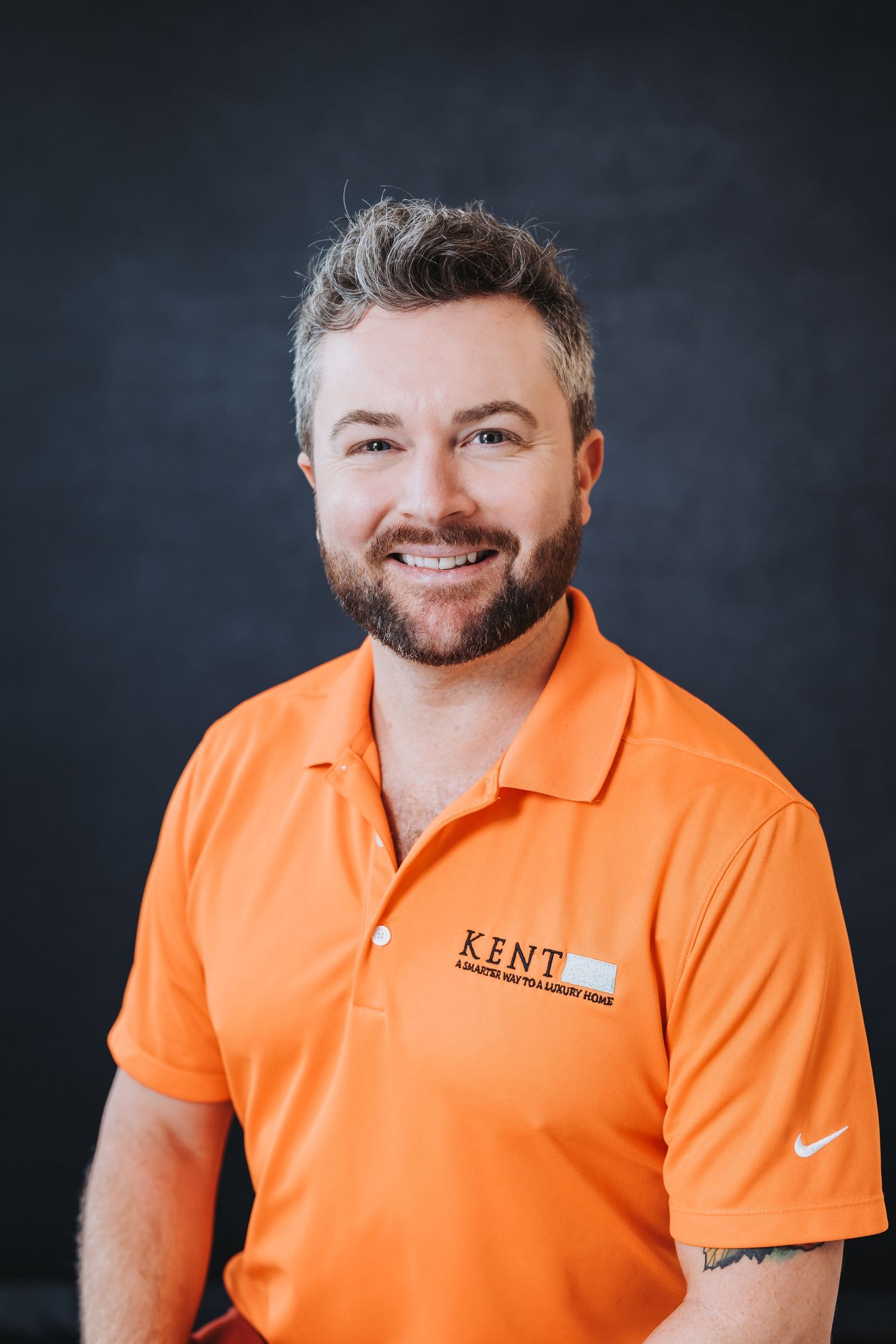 Smiling man with beard wearing an orange Kent polo shirt posed against a dark gray background.