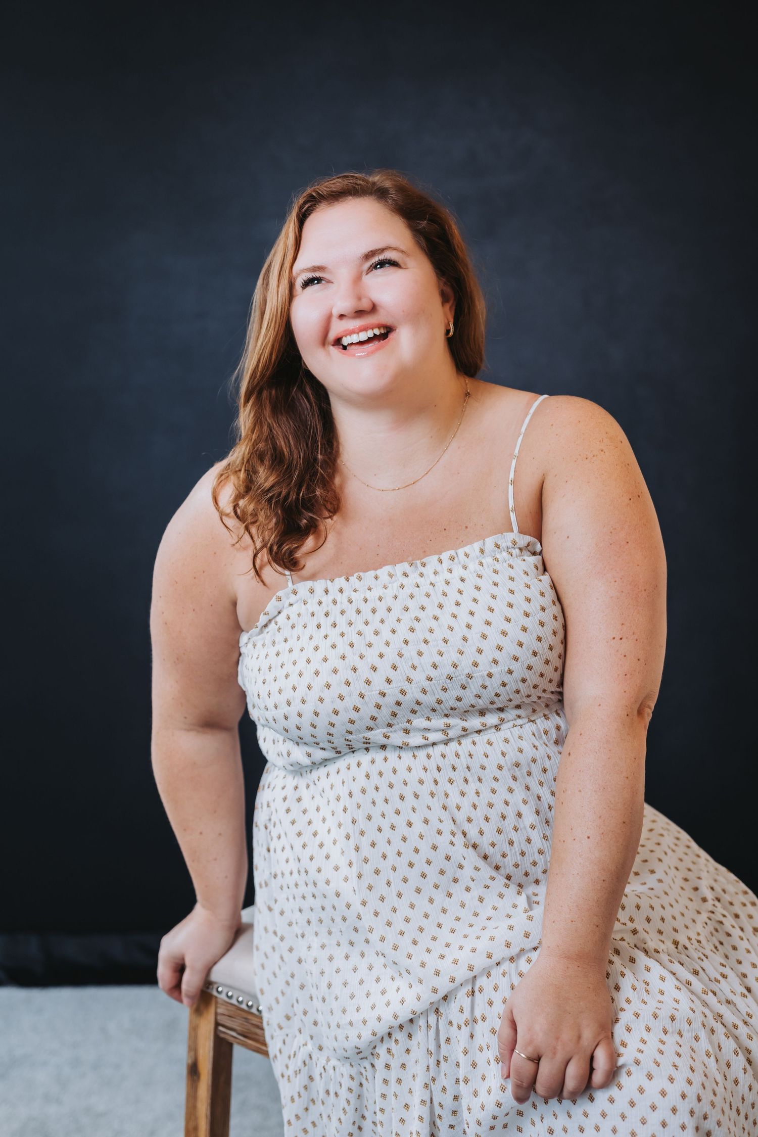 Smiling young woman in a white polka dot sundress sitting on a wooden chair against a dark gray background.
