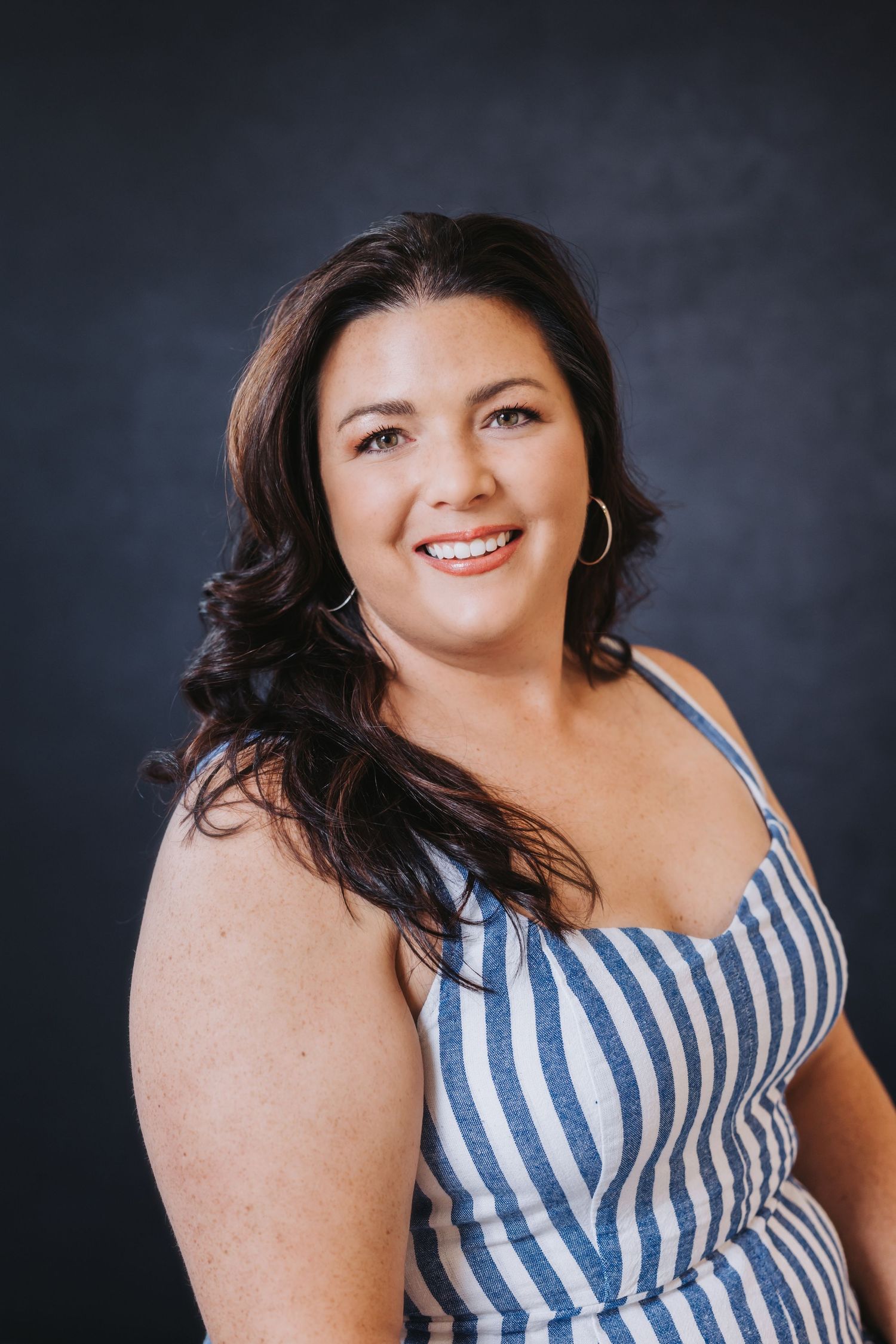 Smiling brunette woman in striped sundress with gold hoop earrings poses against dark gray studio background.