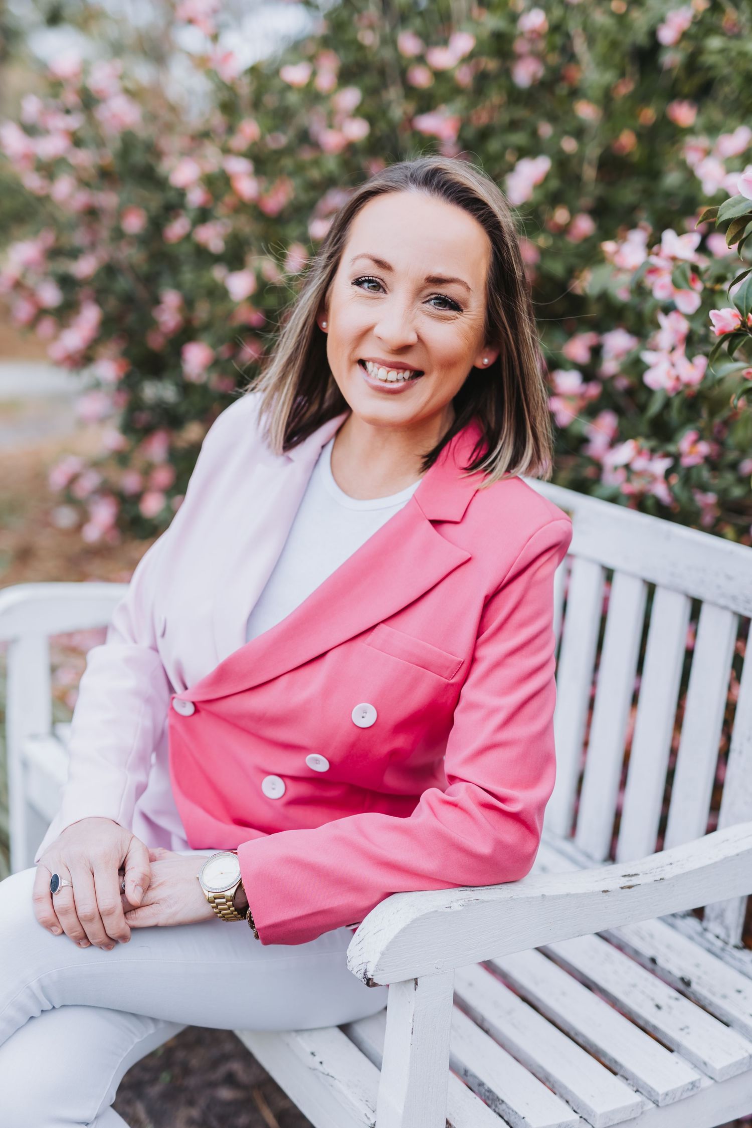 Smiling woman in pink blazer sitting on white bench surrounded by blooming pink flowers outdoors.