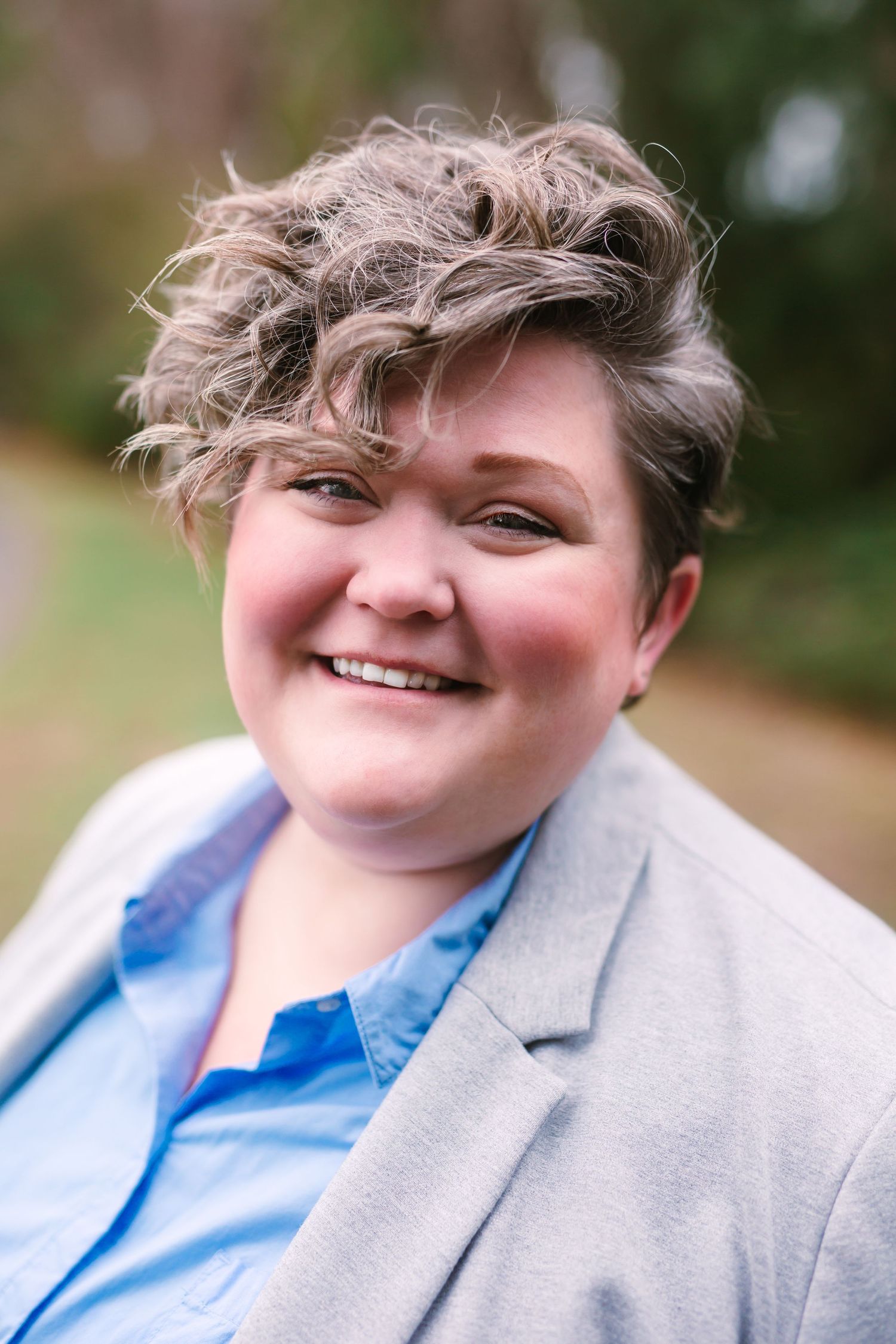 Smiling person with short wavy hair wearing a blue shirt and gray blazer outdoors.