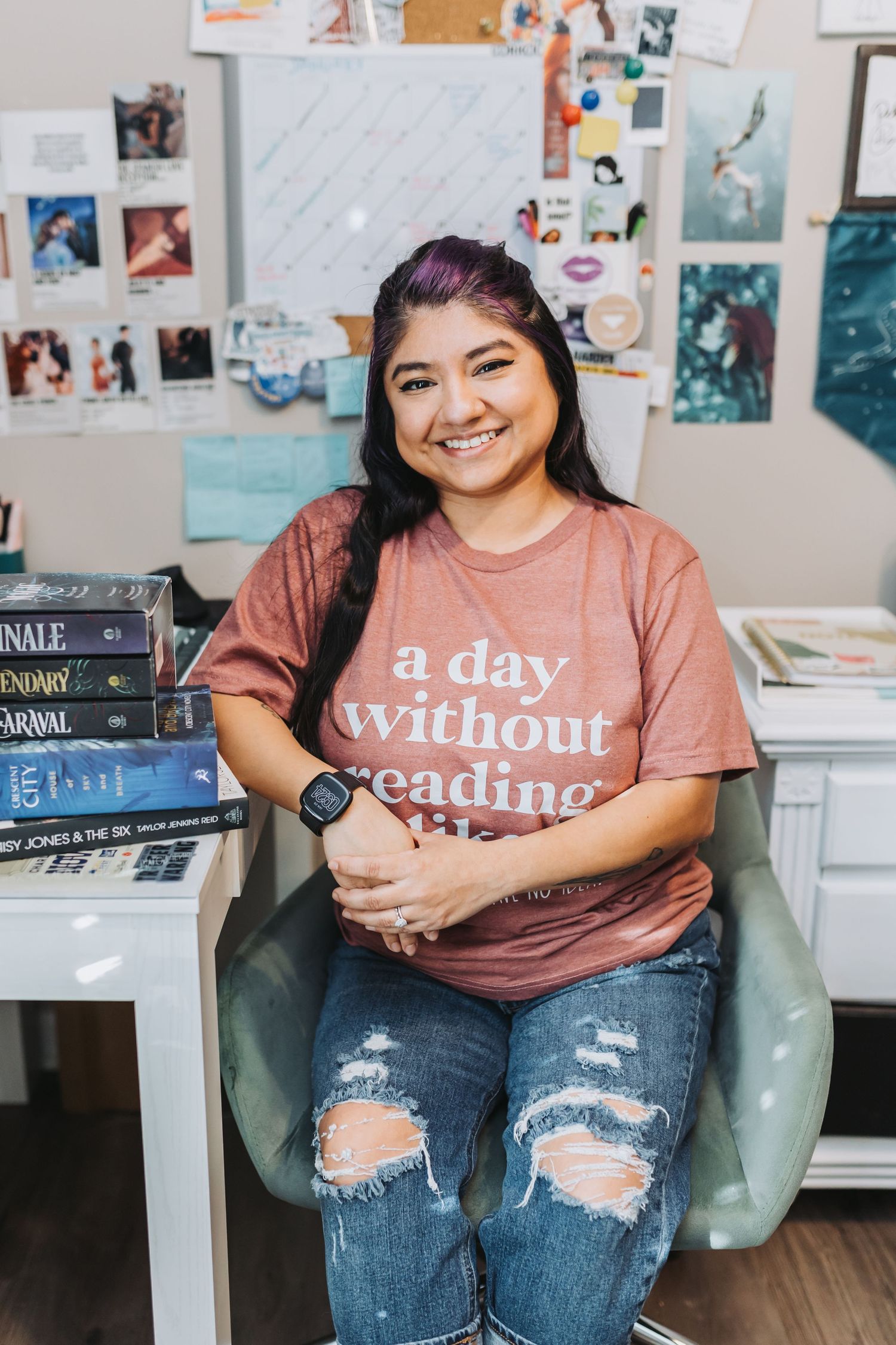 Smiling young woman in ripped jeans and a pink reading t-shirt sits in a chair surrounded by books and a mood board.
