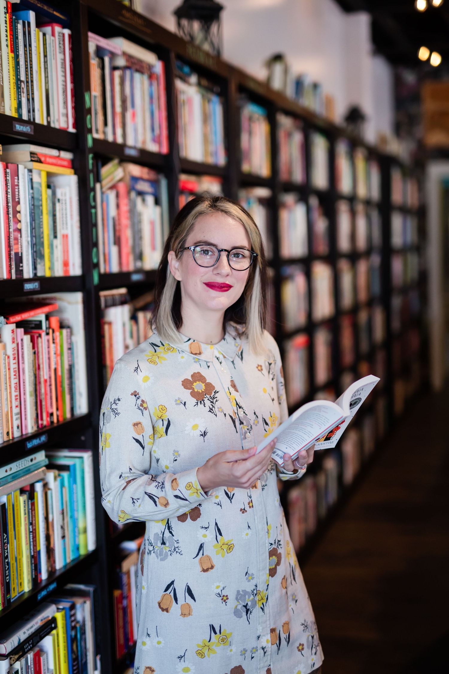 Woman in floral dress holding open book smiling in library with colorful bookshelves.