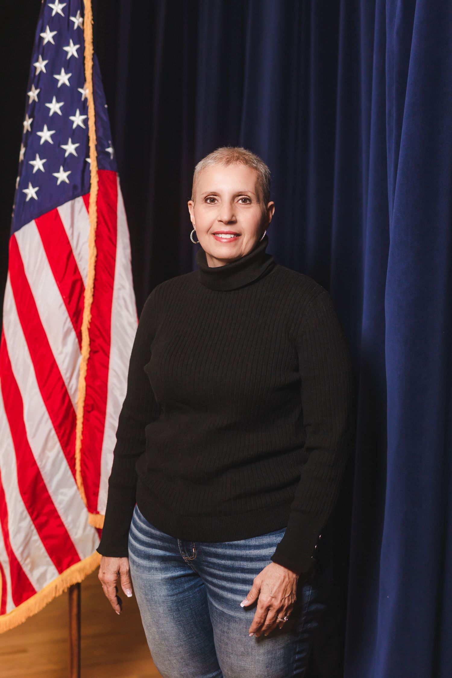 Woman in black turtleneck and jeans smiling beside an American flag against a blue curtain backdrop.