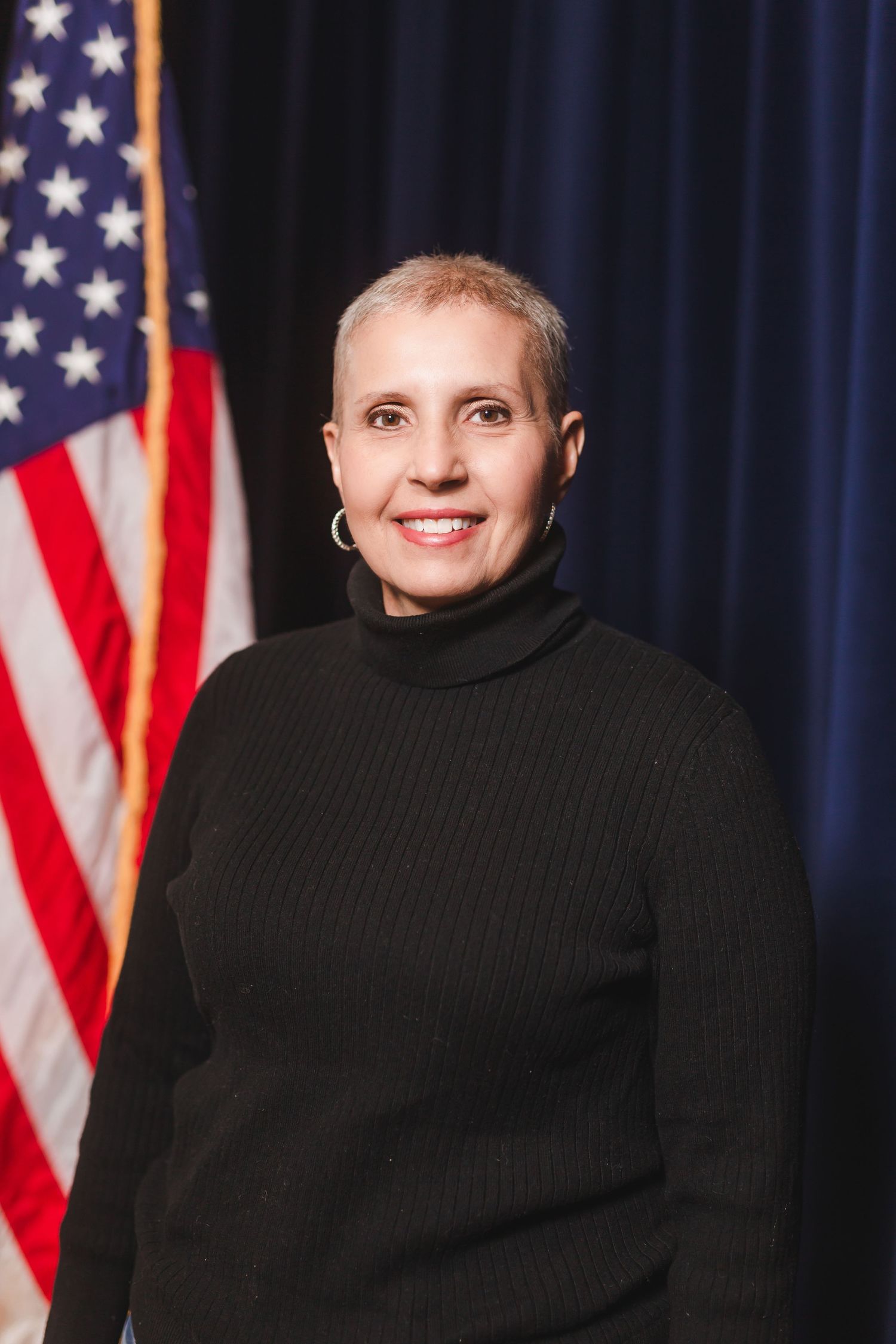 Woman in black turtleneck sweater smiling in front of American flag, professional portrait photo.