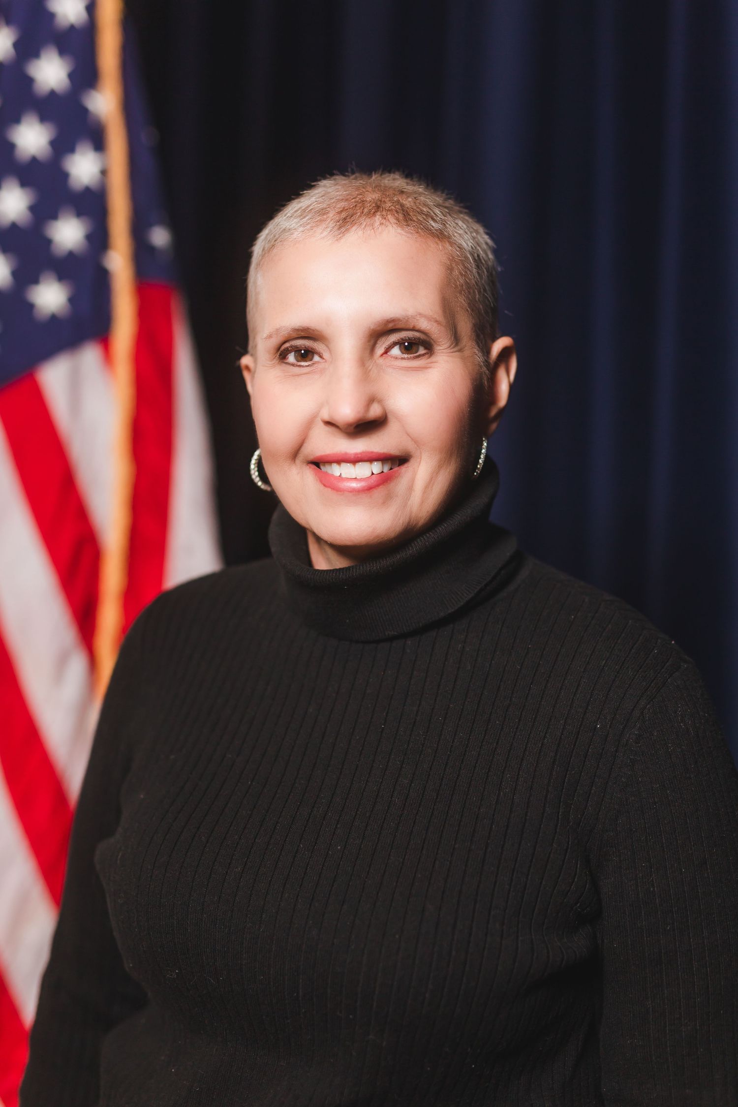 Woman with short blonde hair smiling in black turtleneck sweater posed before American flag.