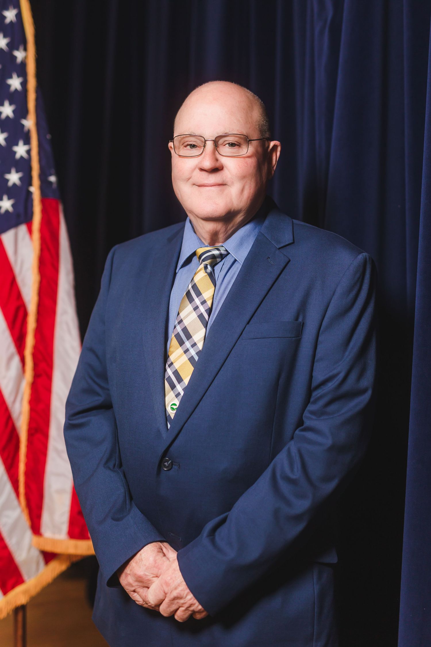 Professional man in blue suit and plaid tie poses before American flag for formal portrait photo.
