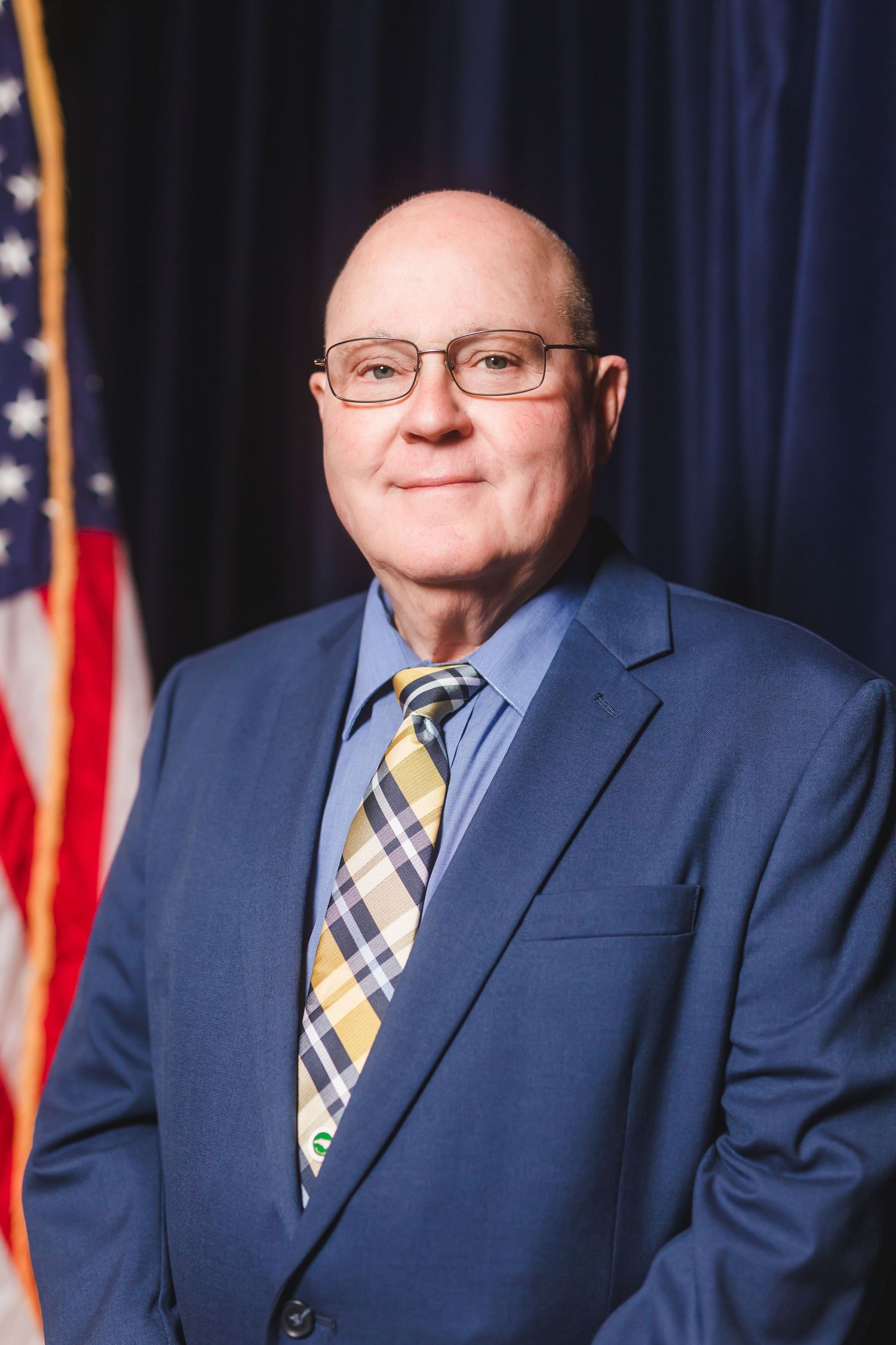 Bald man in blue suit and plaid tie smiling before American flag in professional headshot photo.