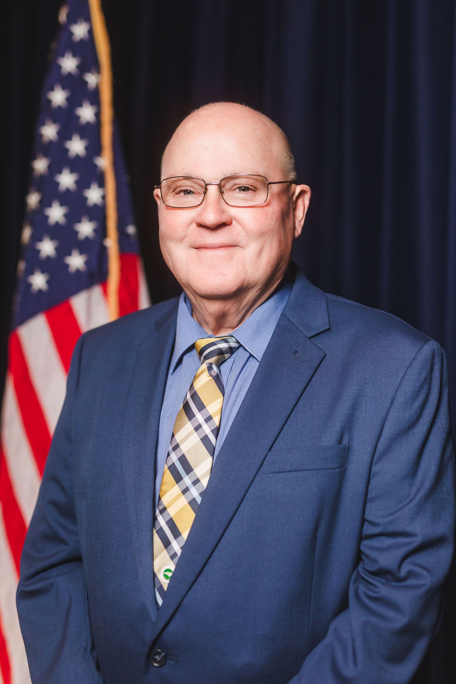 Bald man in blue suit and plaid tie smiling before American flag backdrop.