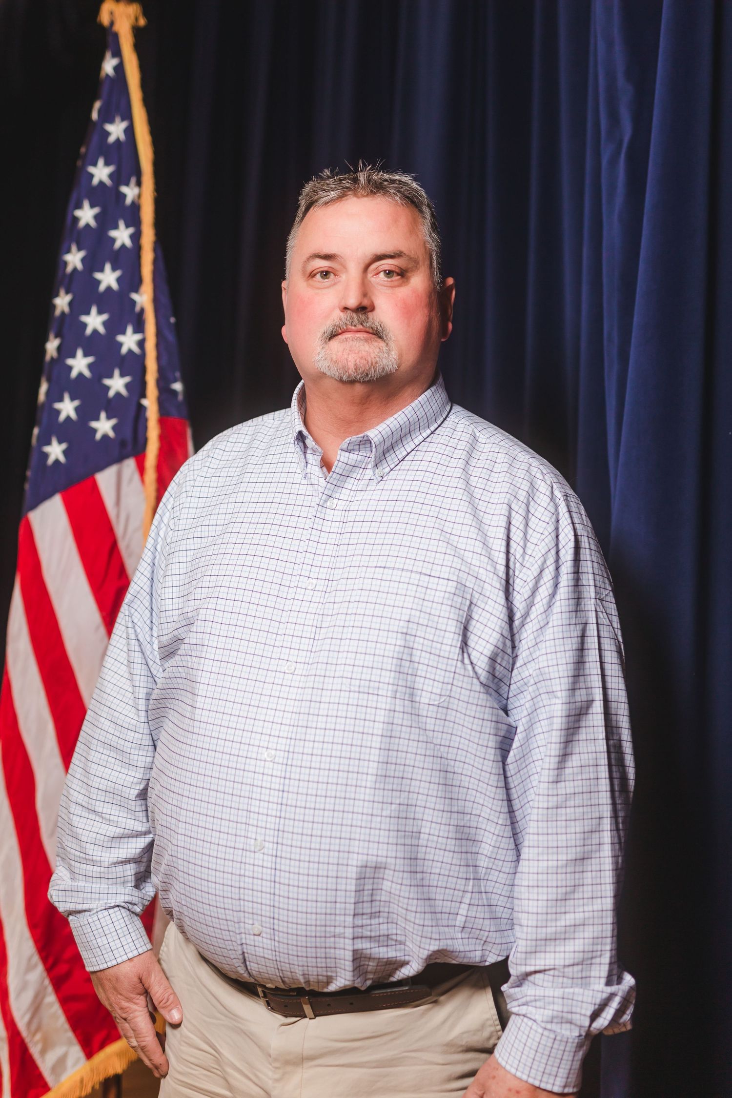 Middle-aged man in a plaid shirt stands before an American flag against a dark blue backdrop.