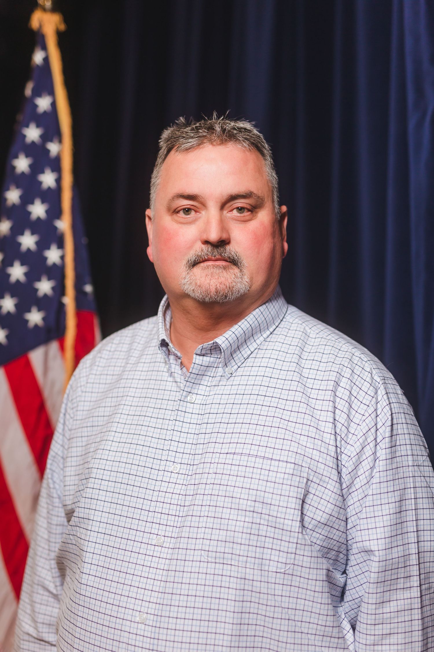 Middle-aged man with goatee in checkered shirt standing before American flag.