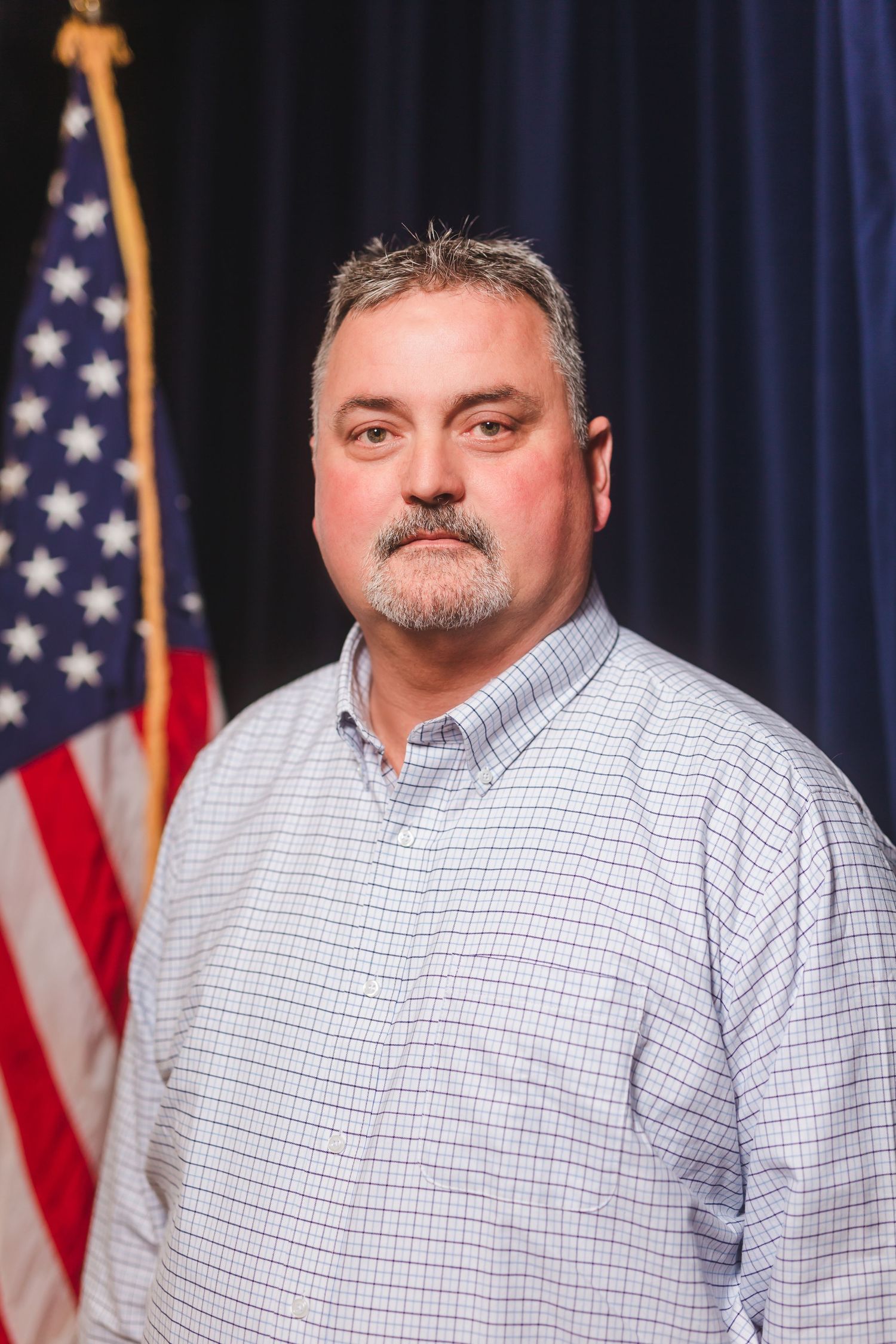 Middle-aged man with gray hair and mustache poses in formal portrait with American flag backdrop.