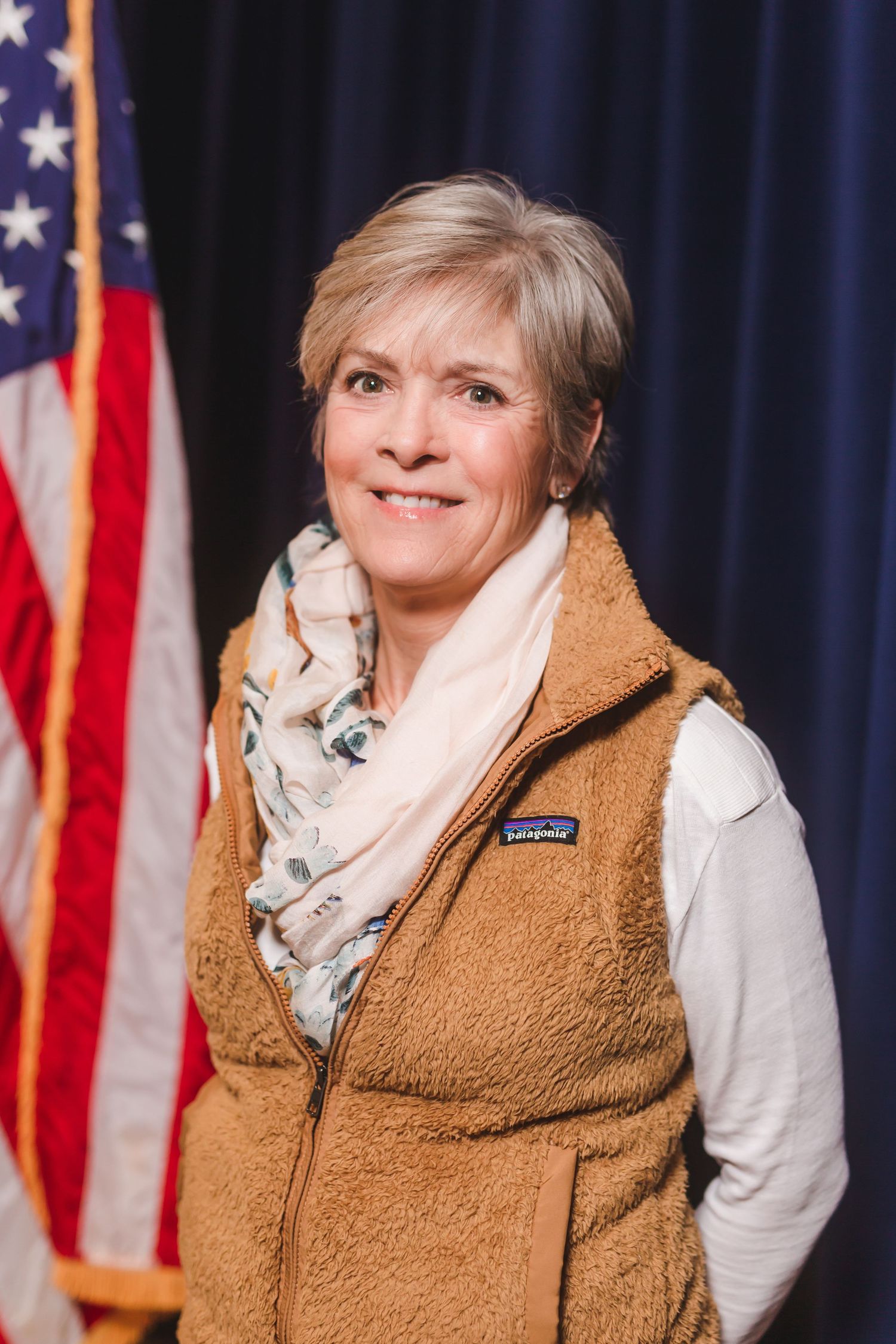 Smiling woman with short gray hair wearing a Patagonia vest and floral scarf standing near an American flag.