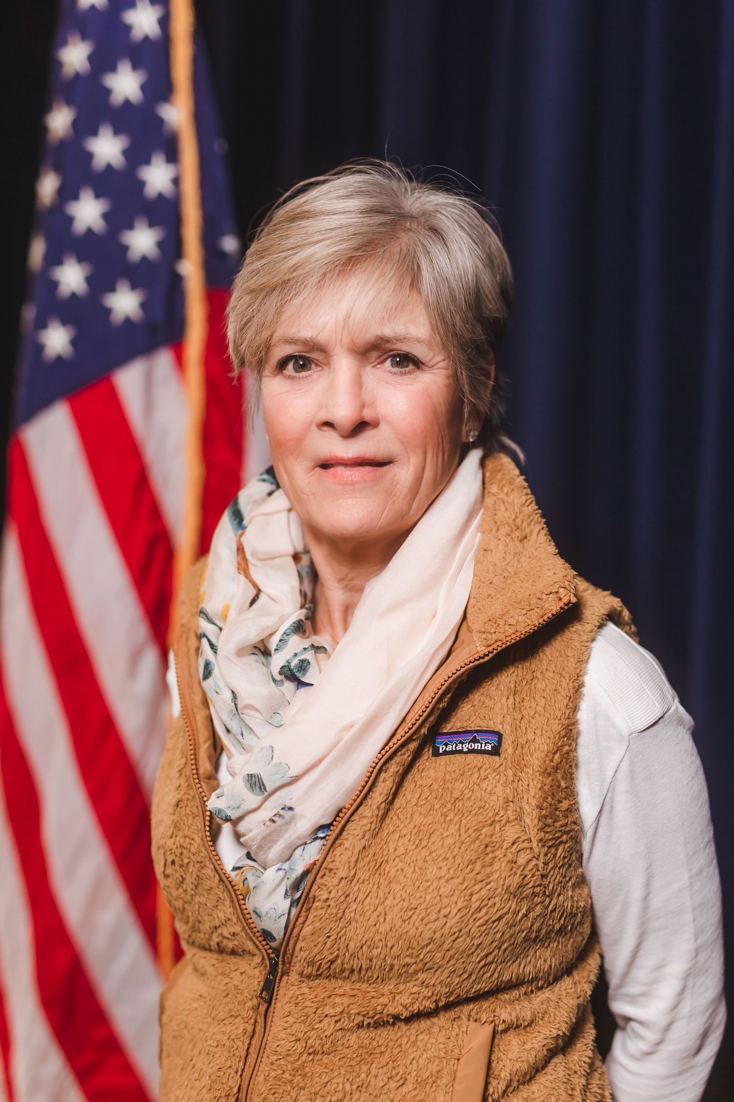 Woman with short gray hair wearing Patagonia vest and scarf before American flag.