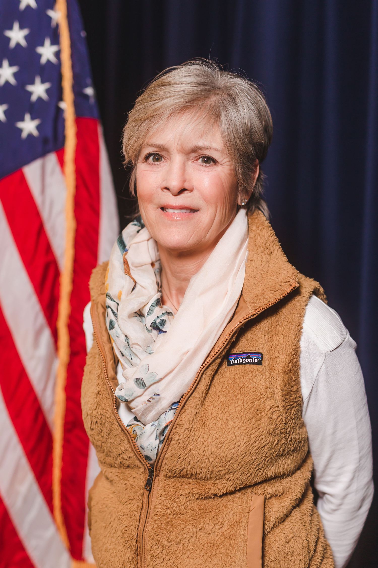 Woman with short gray hair wearing a Patagonia vest and floral scarf, posing in front of an American flag.