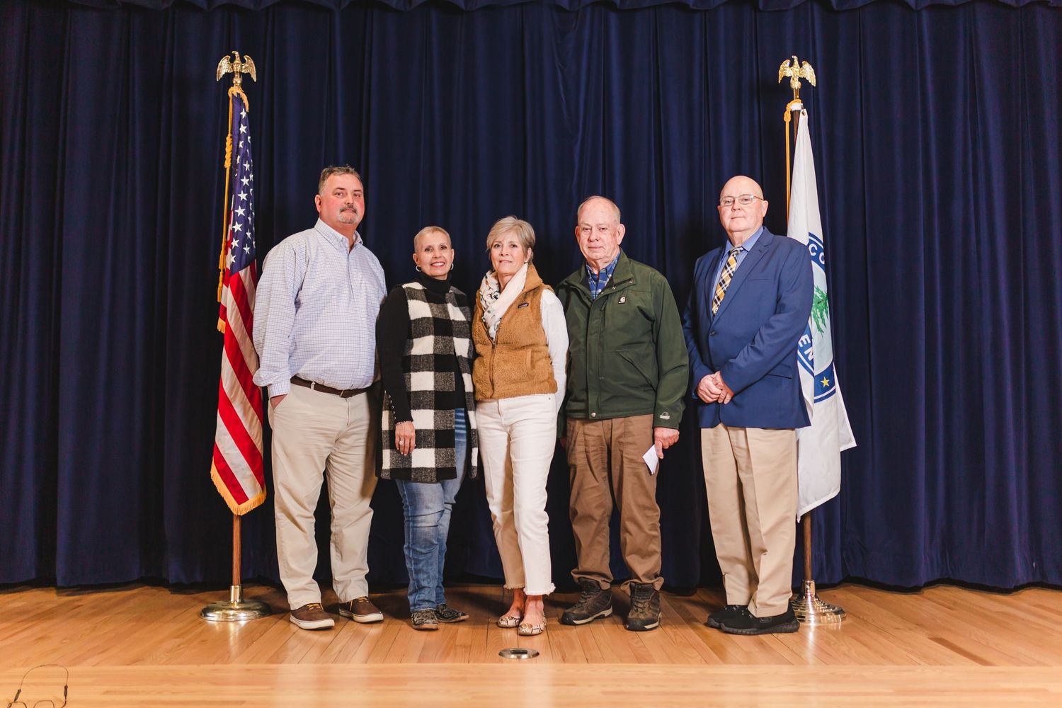 Five adults posing together on a stage between American and state flags at a formal recognition ceremony.