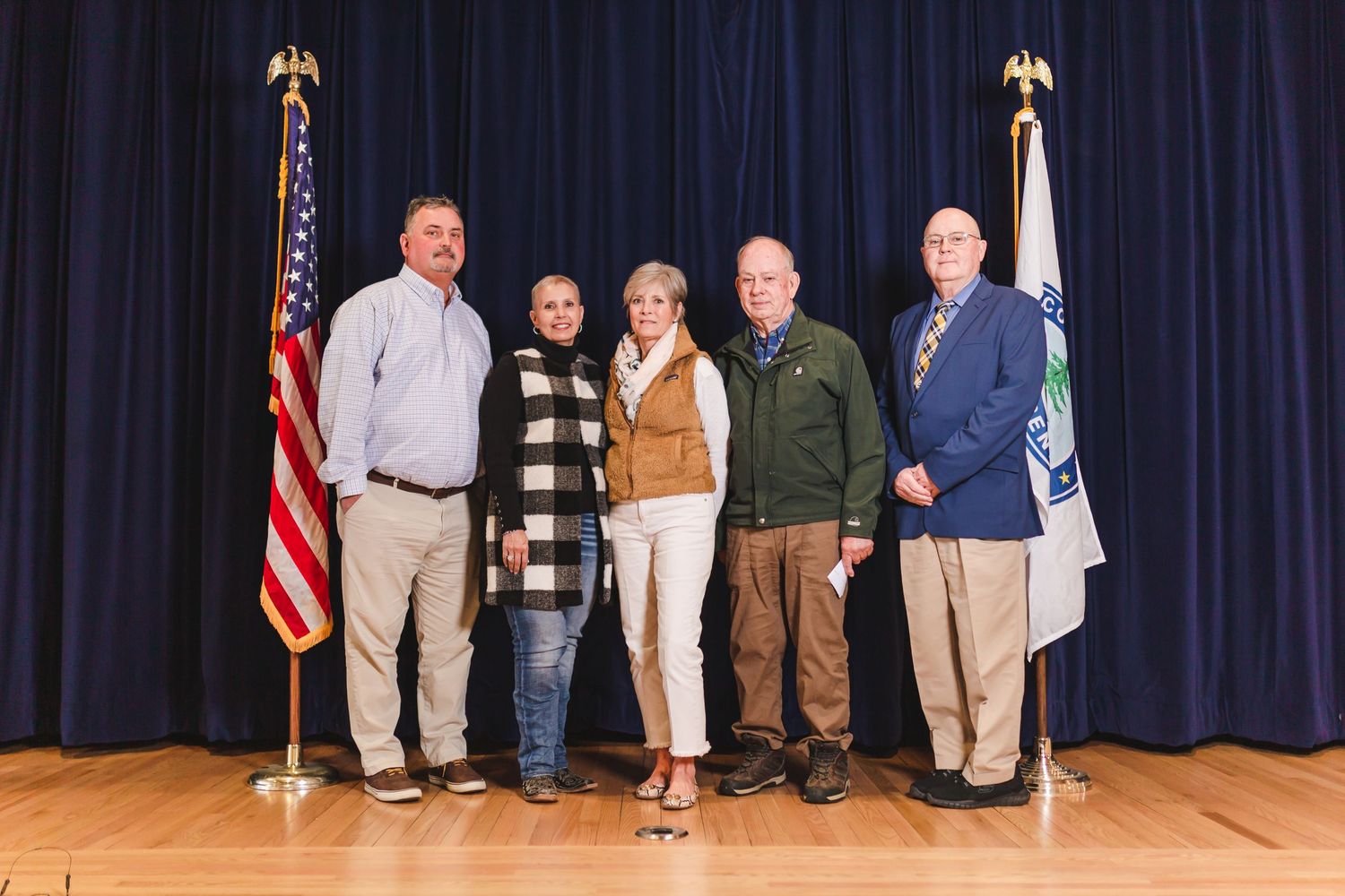 Five adults pose together on a wooden stage between an American flag and a state flag at a formal ceremony.