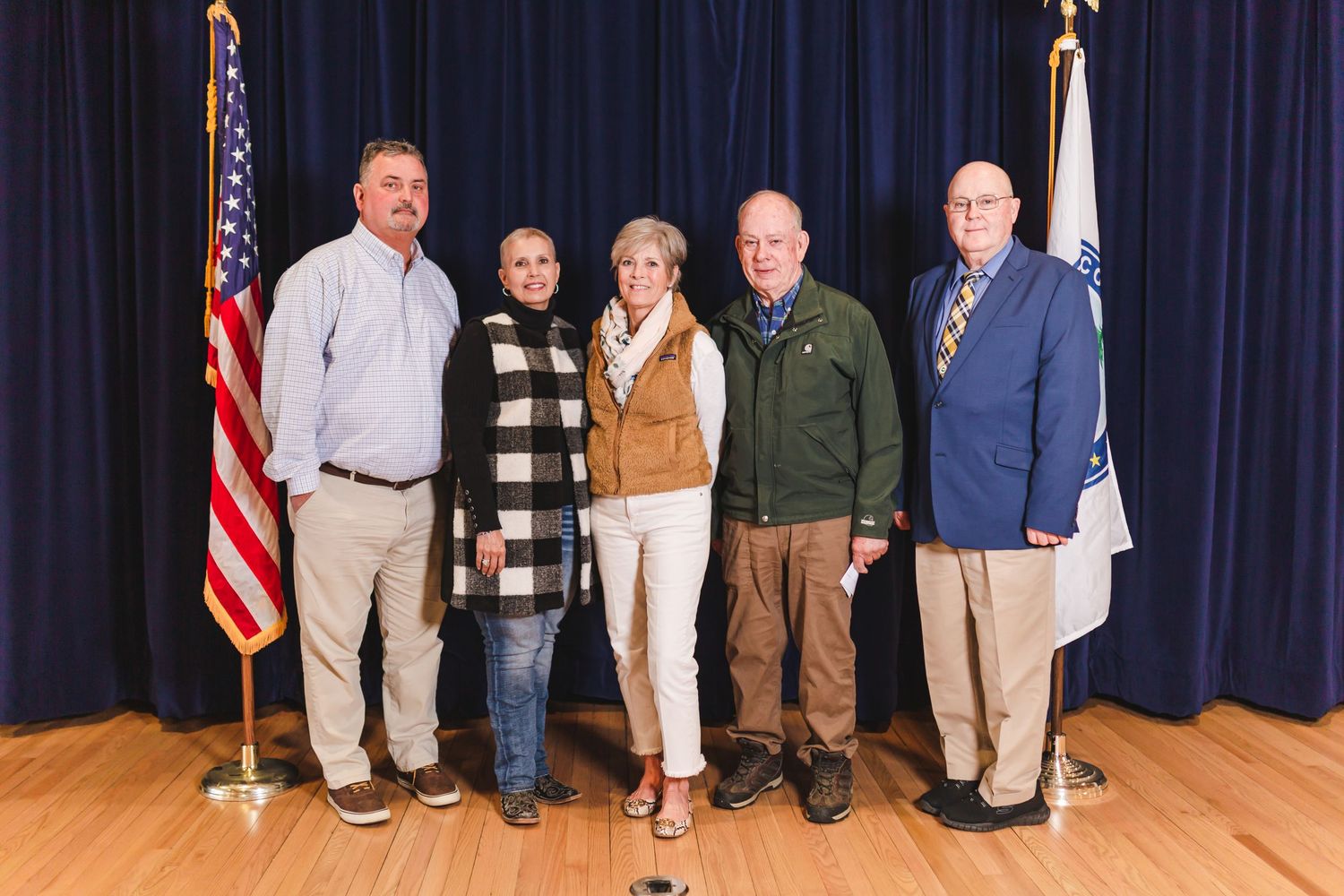 Group of five officials posing together on stage with American flag.