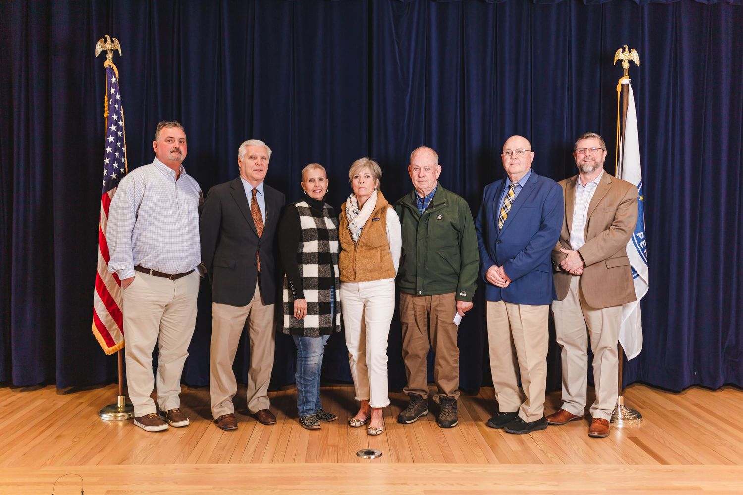 Seven adults posing together on a stage with American flags, at a formal group photo event in an auditorium.
