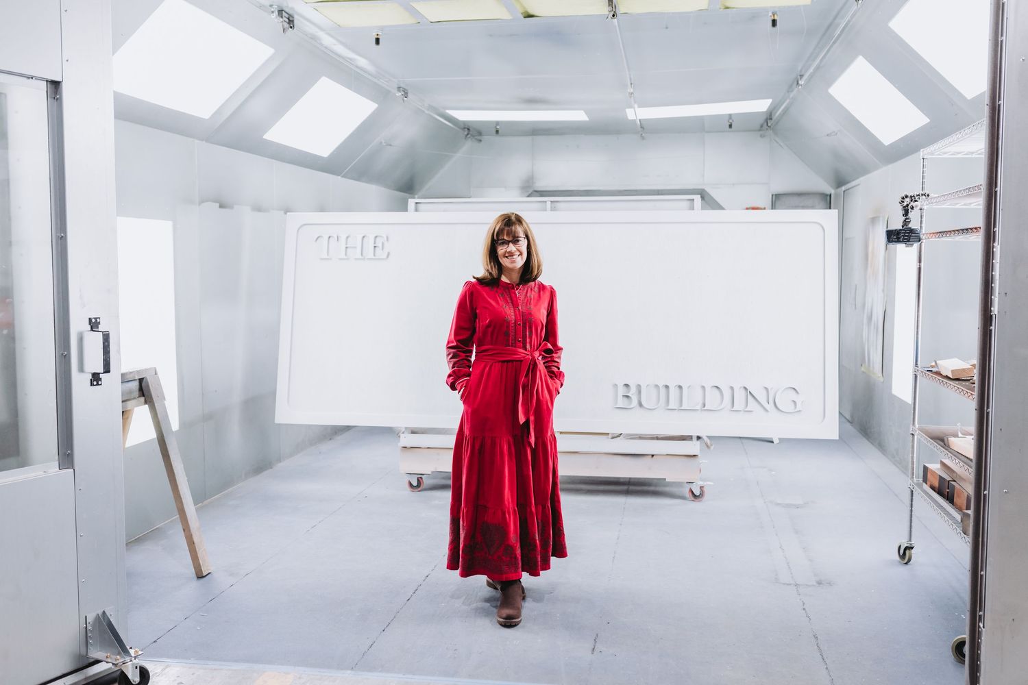 Woman in red dress standing in a white industrial paint booth with 'The Building' text on the wall.