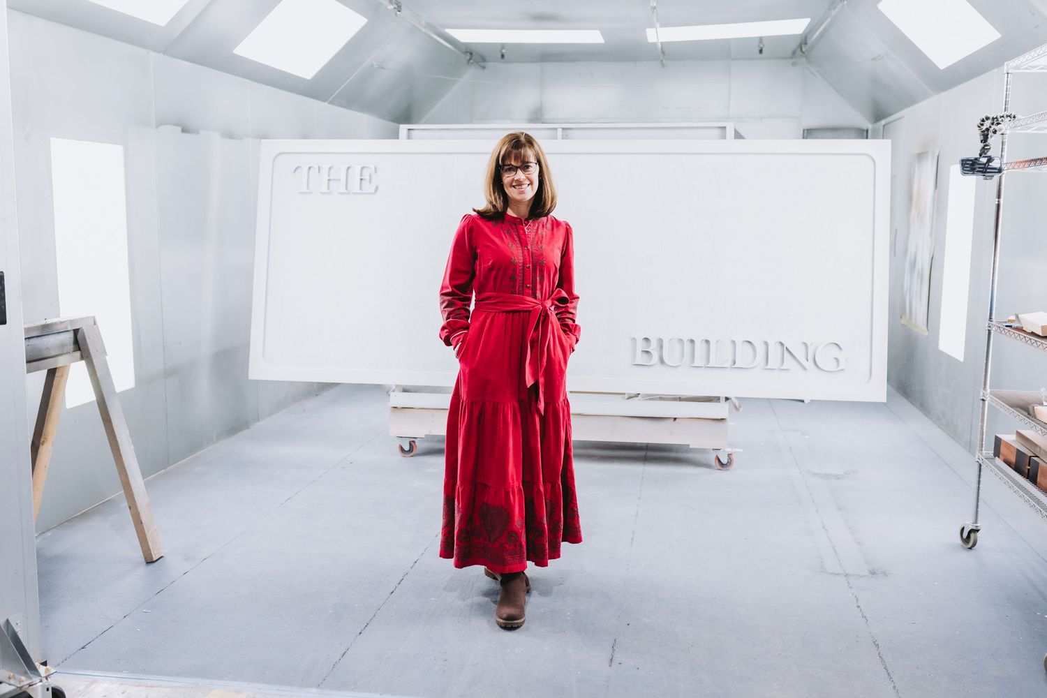 Woman in red maxi dress standing in front of 'The Building' sign in a bright white studio space.