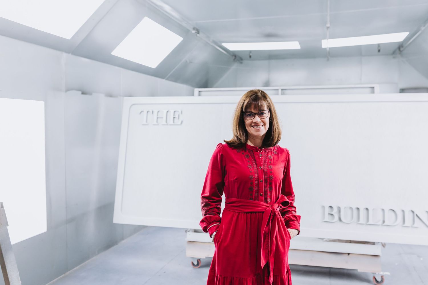 Woman in red dress stands confidently in a white studio space with 'The Building' text backdrop.