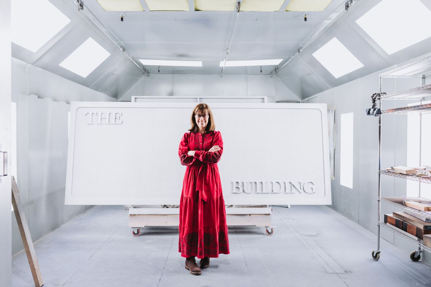 Woman in red dress stands before a white sign reading THE BUILDING in a bright white industrial spray booth.