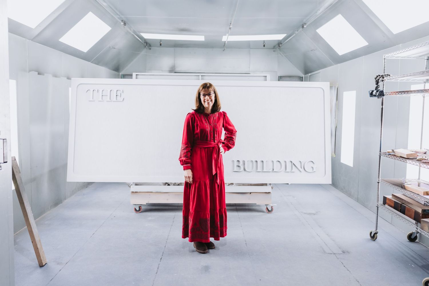 Woman in red dress stands before a white wall sign reading THE BUILDING in a bright studio space.