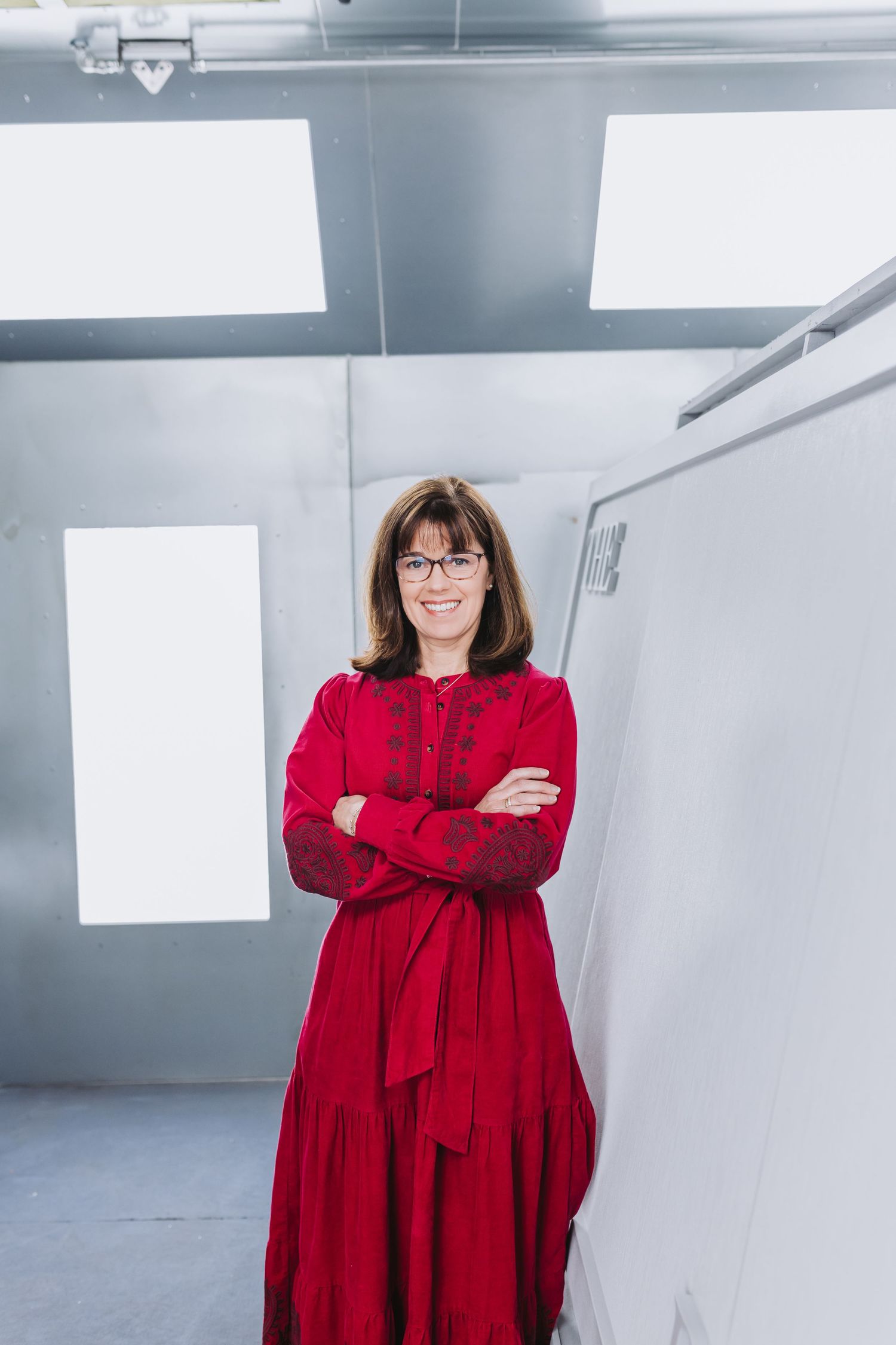 Smiling woman in red dress with arms crossed standing in a modern white studio.