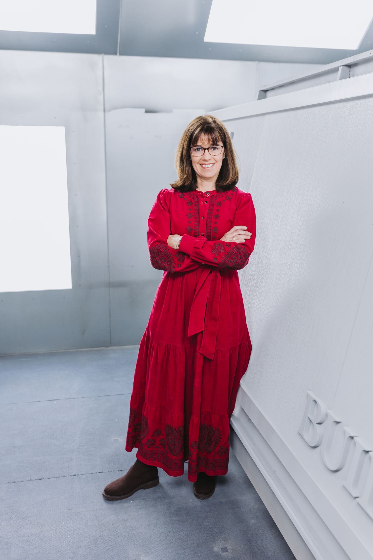Woman in red maxi dress with embroidery, arms crossed, standing confidently in a modern white interior space.