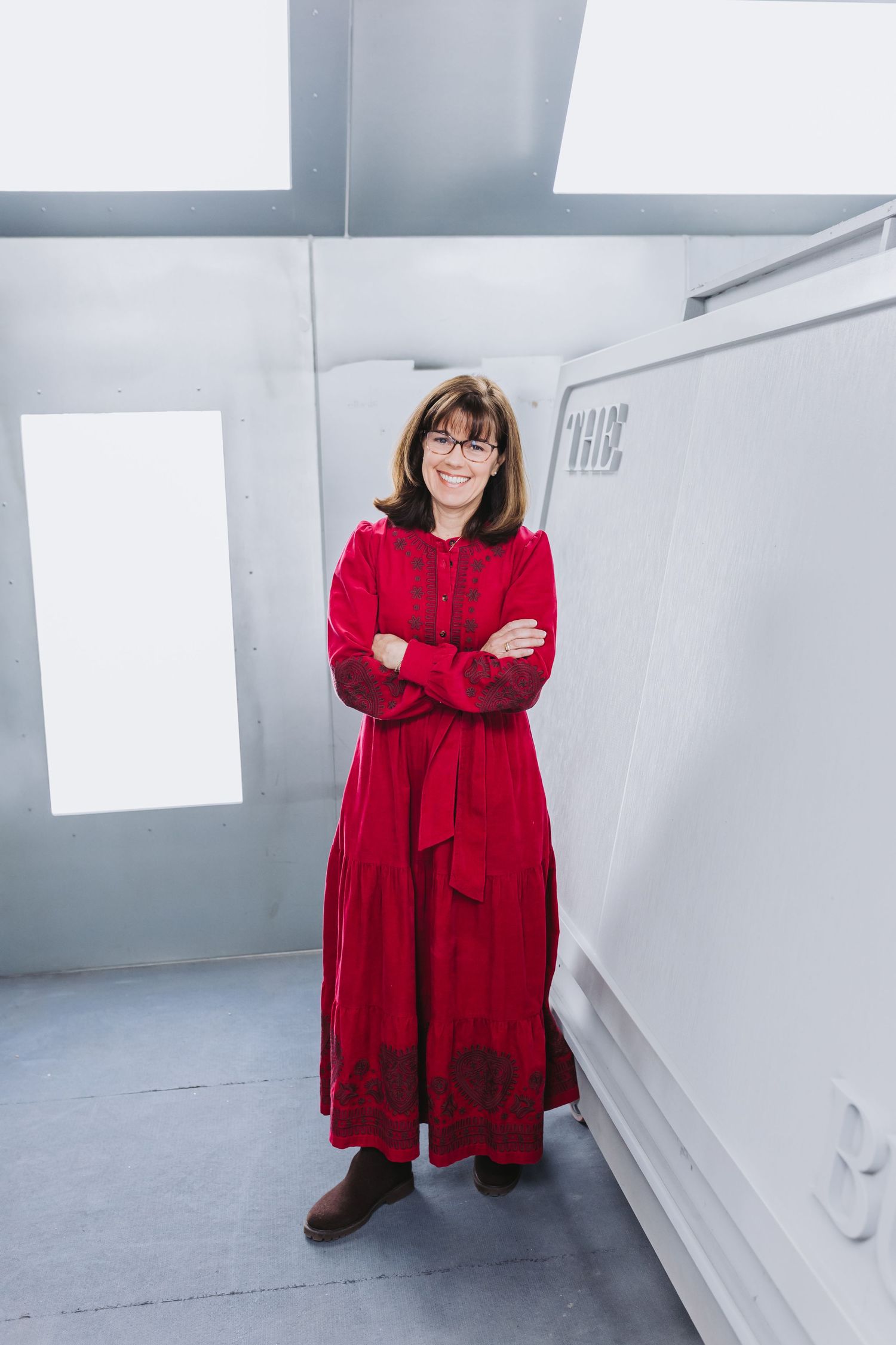 Woman in red dress with glasses stands arms crossed, smiling in a modern gray interior space.