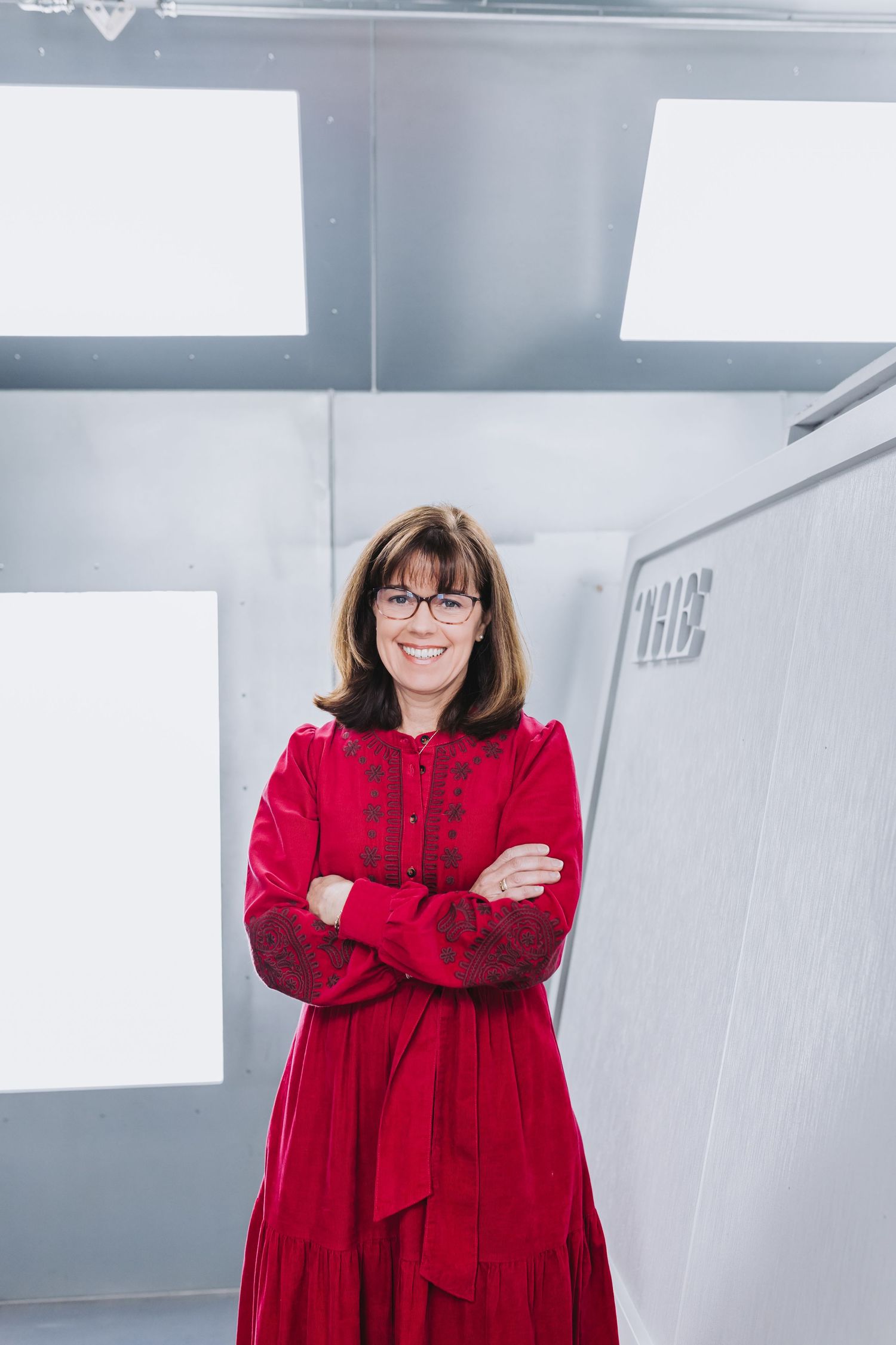 Woman in red dress with glasses smiling, arms crossed, standing in modern silver interior space.