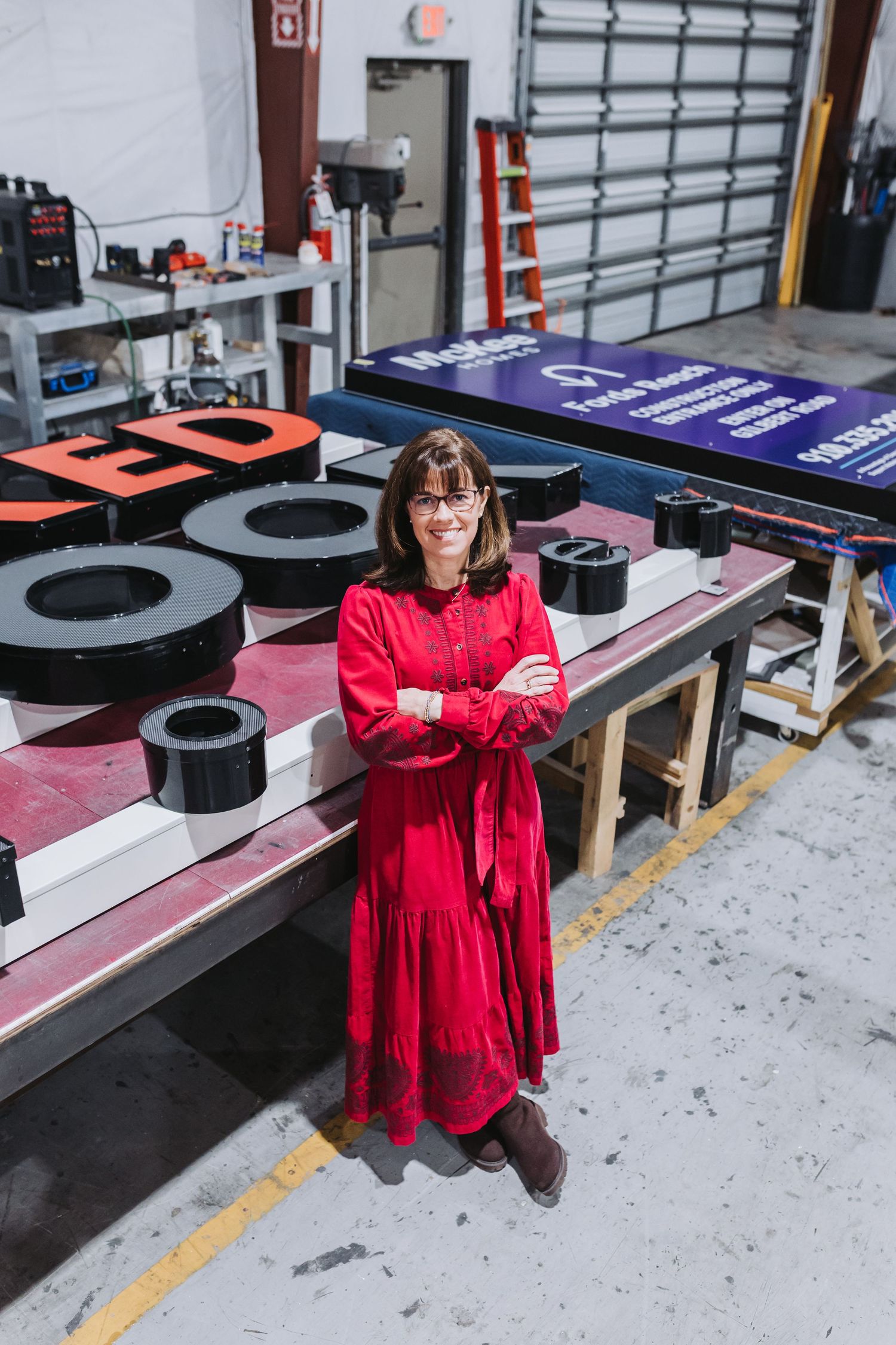Woman in red dress stands smiling with arms crossed in a sign manufacturing workshop with large letters on a worktable.