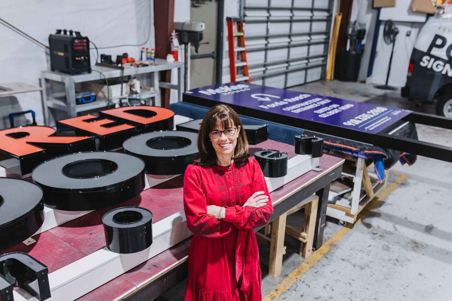 Woman in red dress stands confidently in a sign-making workshop surrounded by large colorful letters and signage.