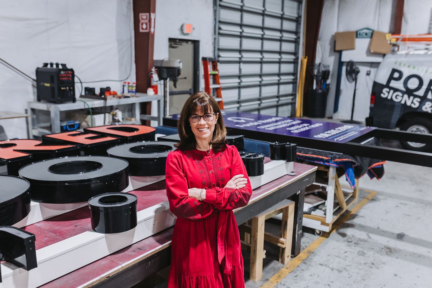 Woman in red dress stands confidently in a sign manufacturing workshop with arms crossed, surrounded by colorful sign materials.