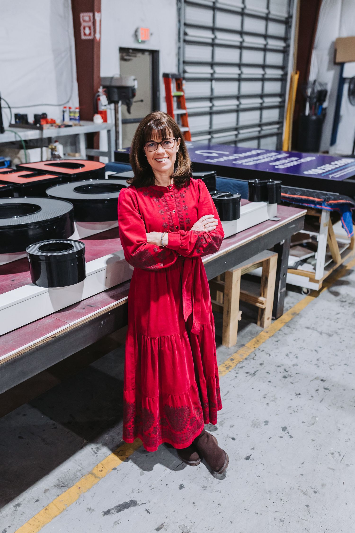 Woman in red dress stands confidently with arms crossed in an industrial workshop setting.