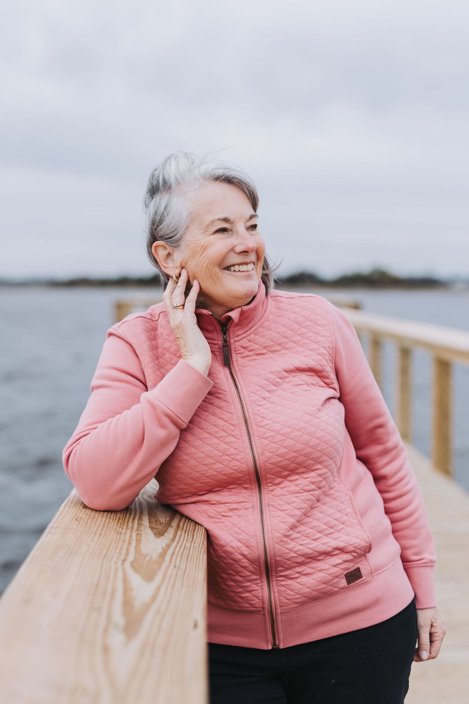 Smiling older woman in pink quilted zip-up jacket leaning on wooden pier railing with water in background.