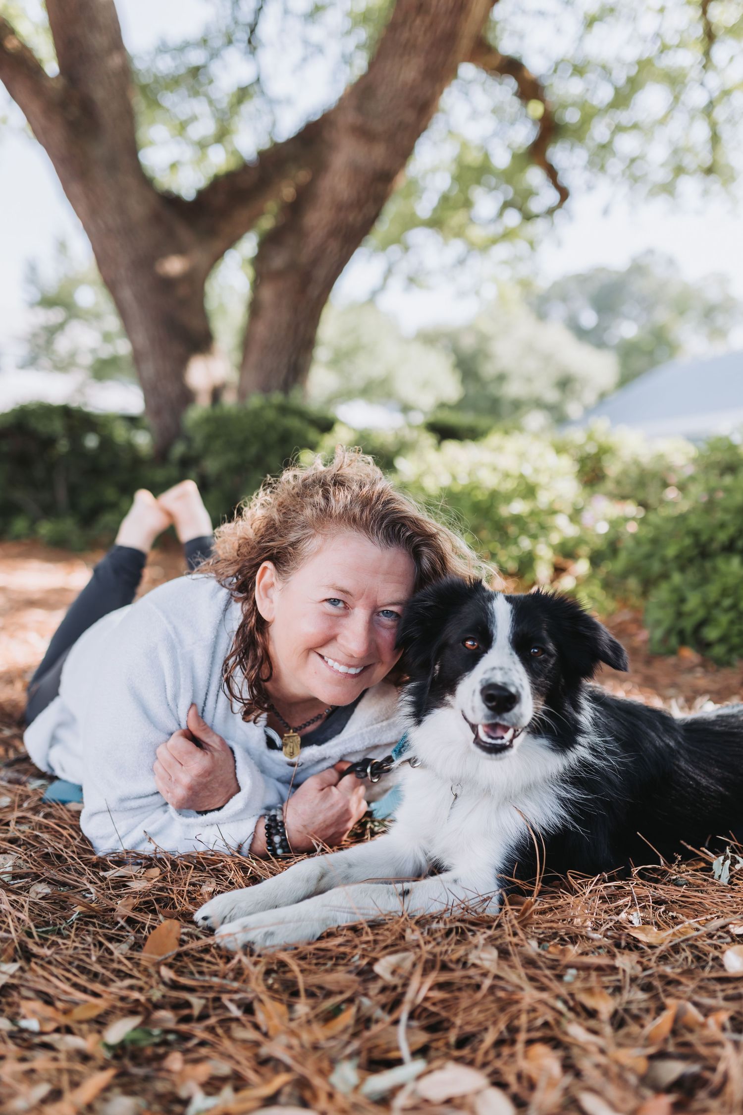 A smiling woman with curly hair lies on fallen leaves with a black and white Border Collie dog under a large tree.