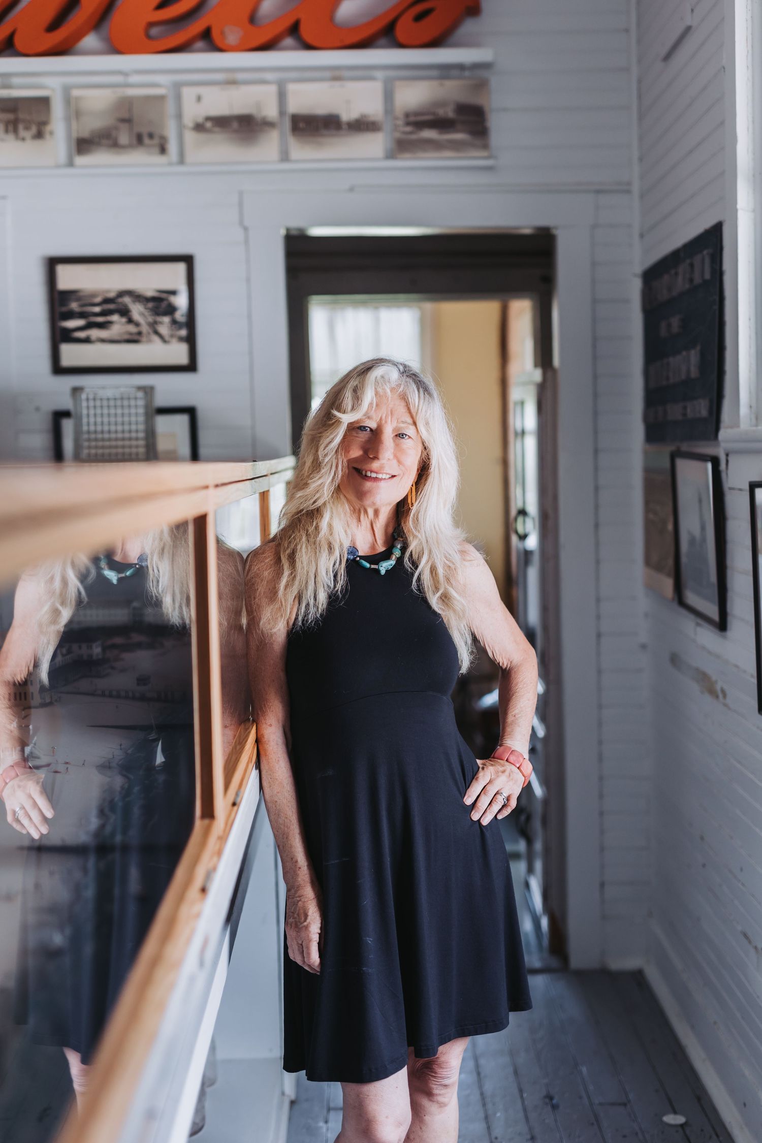 Smiling older woman with long blonde hair wearing a black halter dress and turquoise necklace standing indoors.
