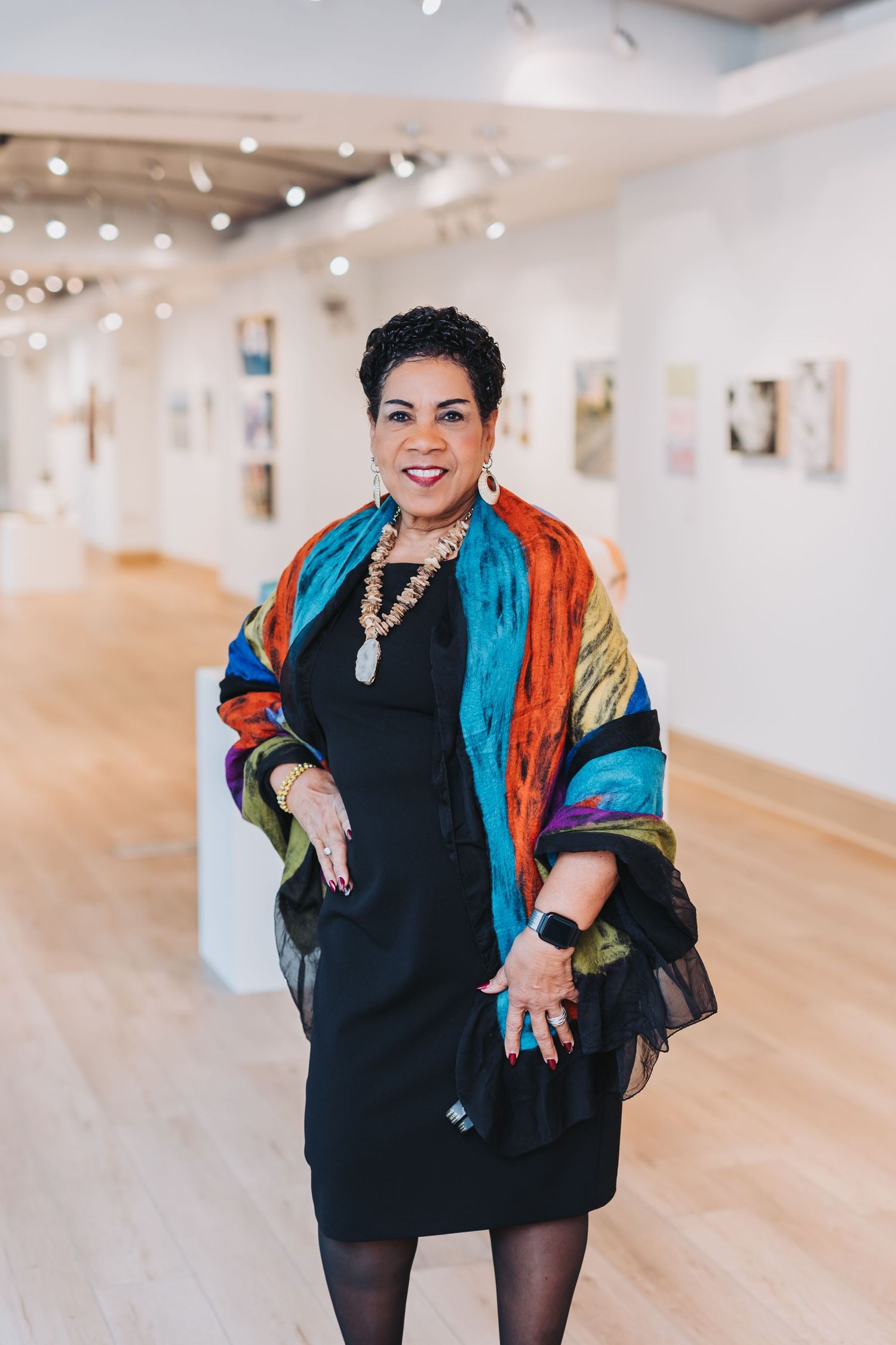 Woman in black dress and colorful rainbow shawl smiling in bright art gallery with wooden floors.