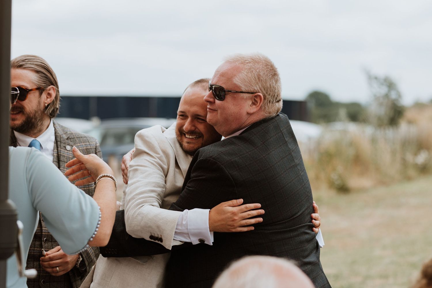 Two men sharing a warm hug at an outdoor wedding celebration, with guests visible in the background.