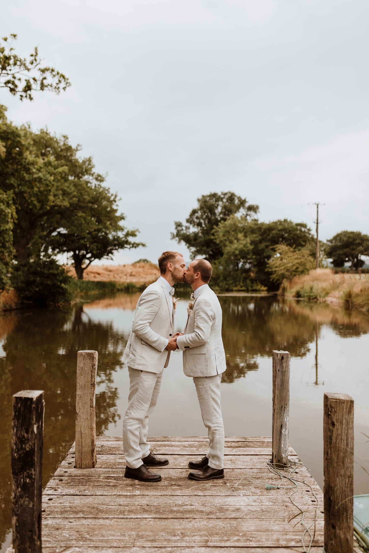 Two grooms share a kiss on a wooden dock overlooking a serene lake on their wedding day at Moat Hall Barns